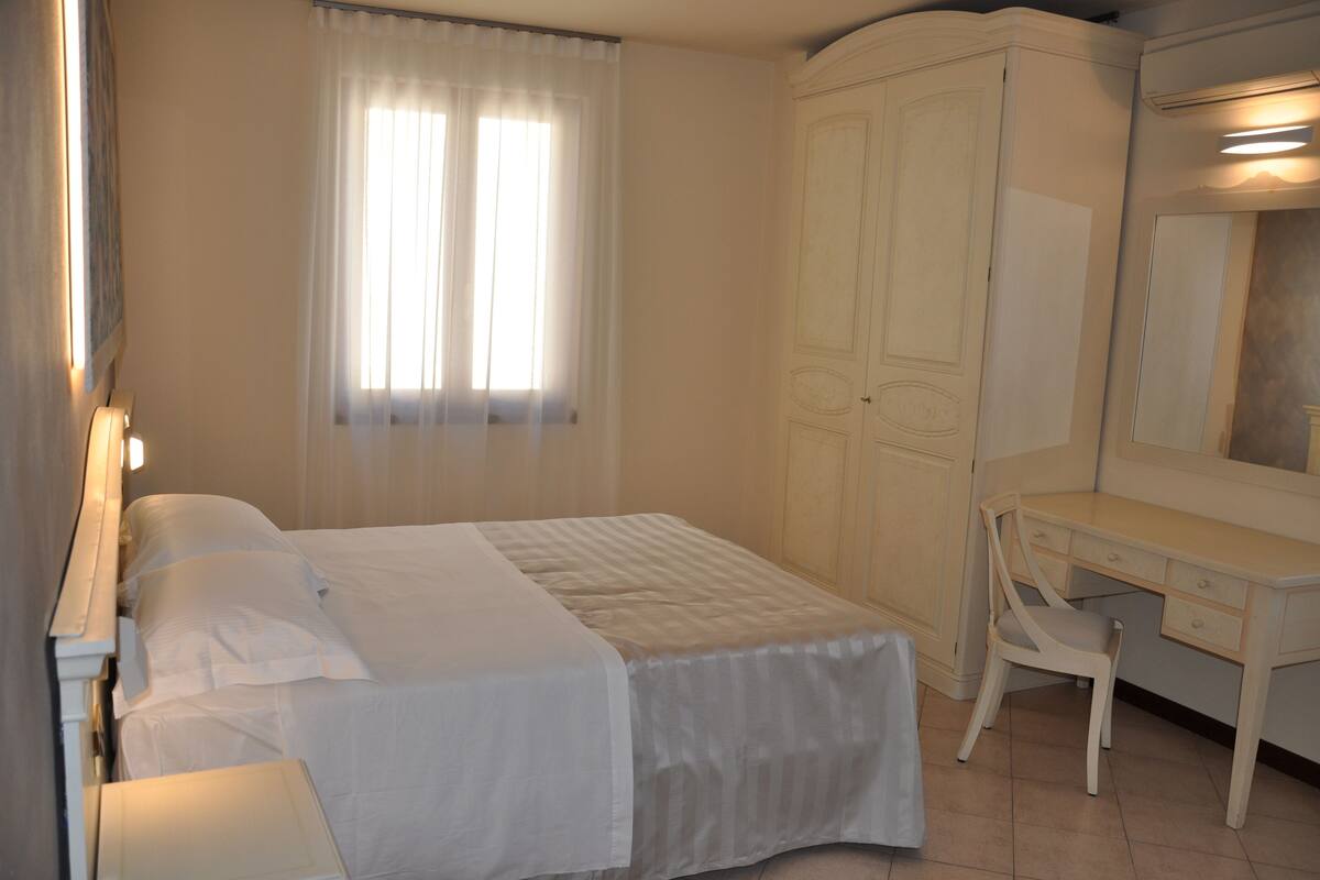 A neatly made bed with white linens, a bedside table with a lamp, a wooden desk with a chair, and a wardrobe with a mirror.