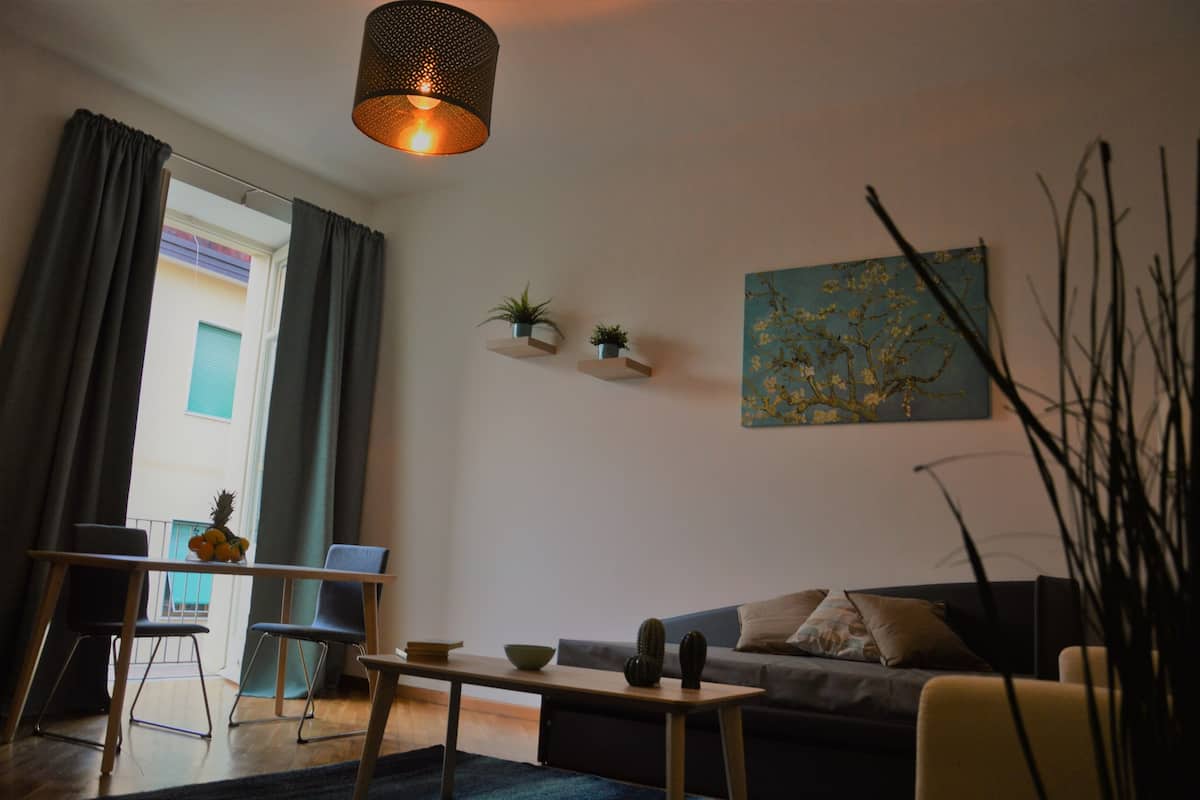 A living room with a sofa, a coffee table, and a dining table with chairs. There are two wall-mounted shelves with plants, a hanging light fixture, and a painting on the wall.