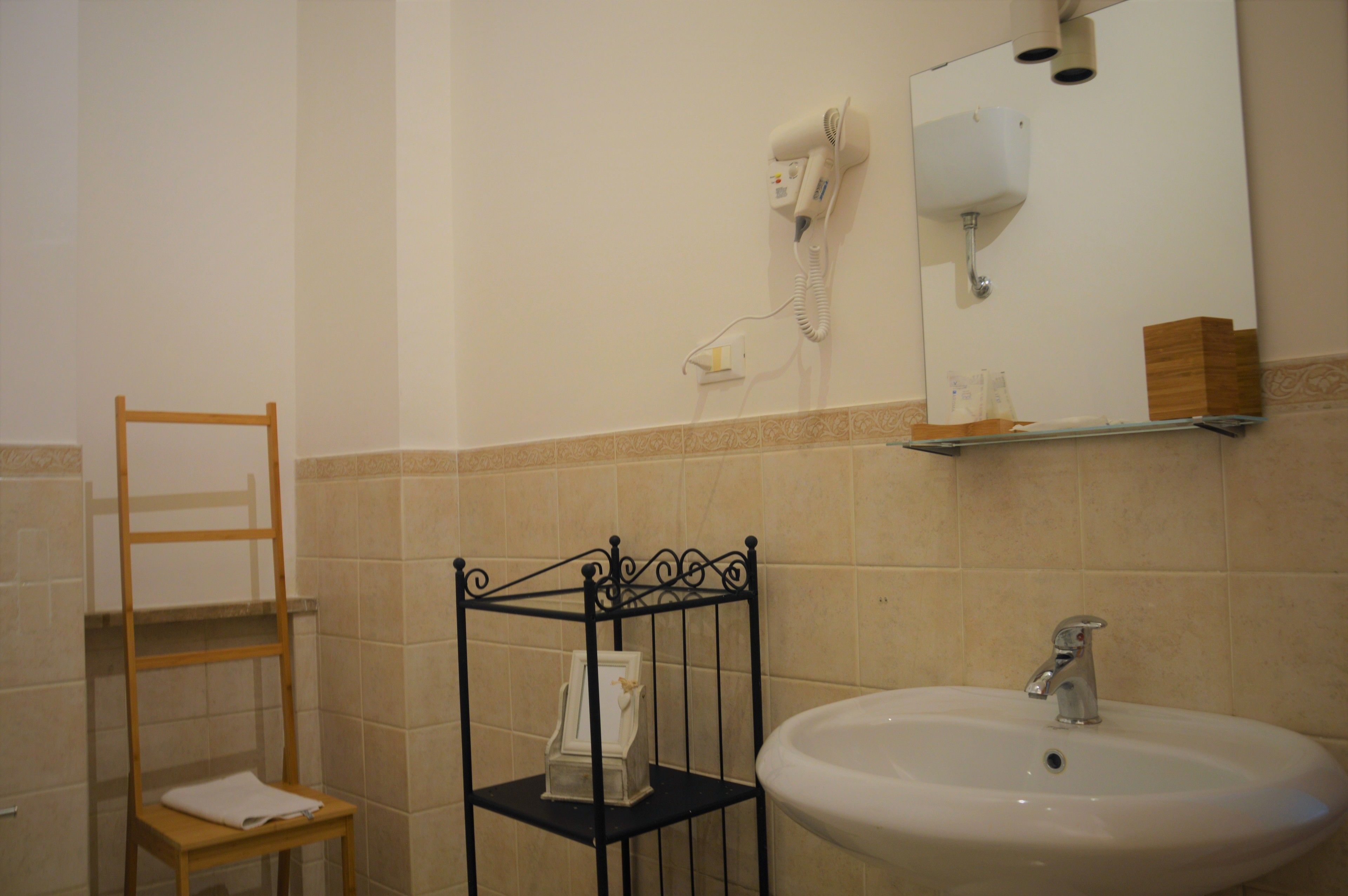 A bathroom with a white sink, a hairdryer mounted on the wall, a shelf with toiletries, and a wooden chair.