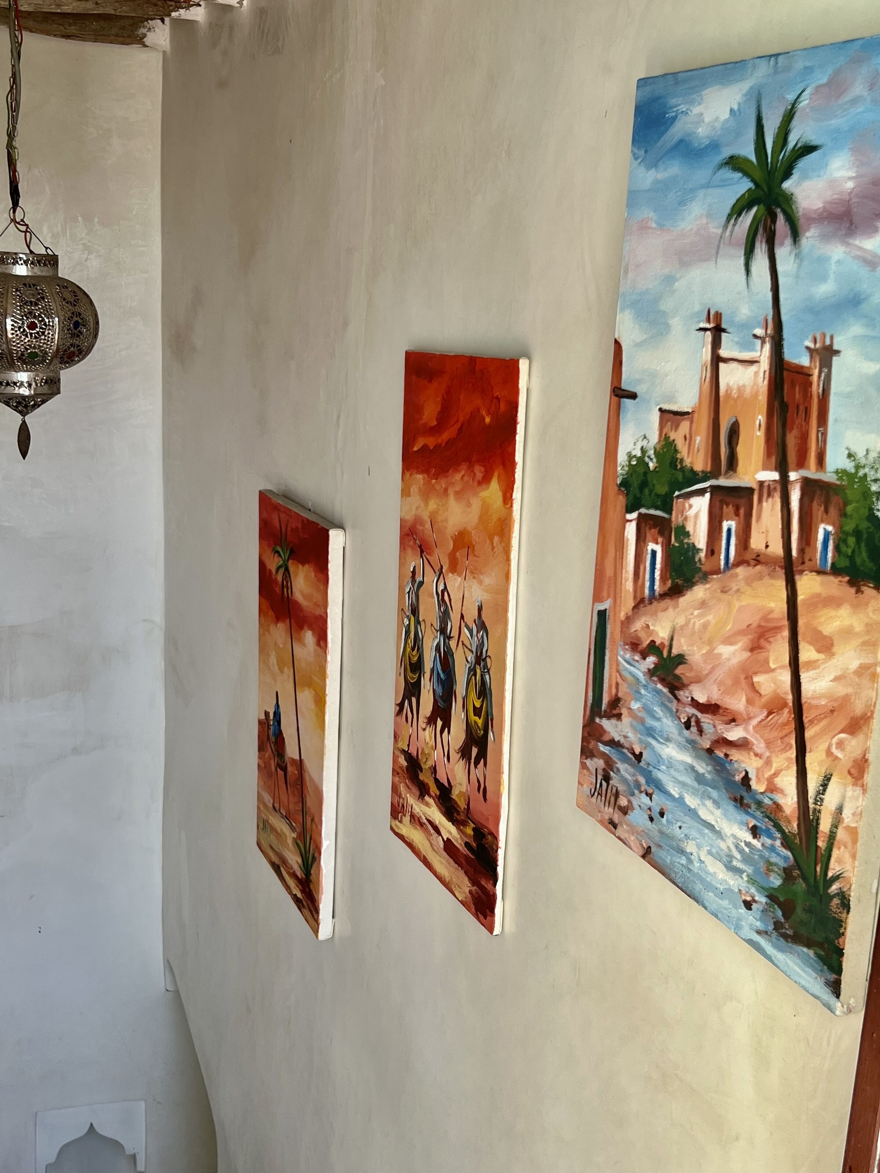 Three paintings on a wall, depicting various scenes including a sunset, a group of people on horses, and a building with a palm tree.