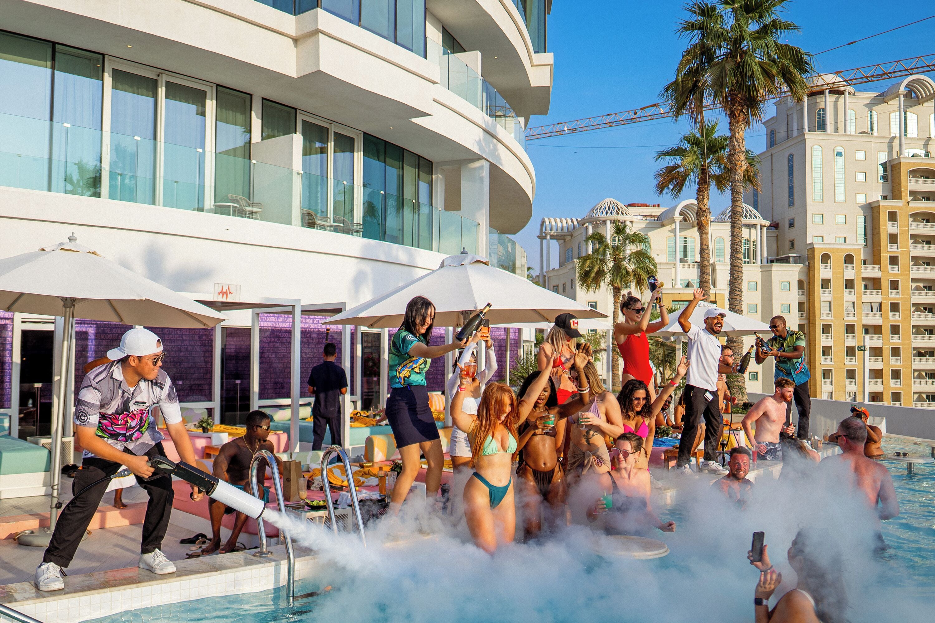 A poolside area with a modern building in the background, a group of people enjoying a pool party, and a person using a water cannon.