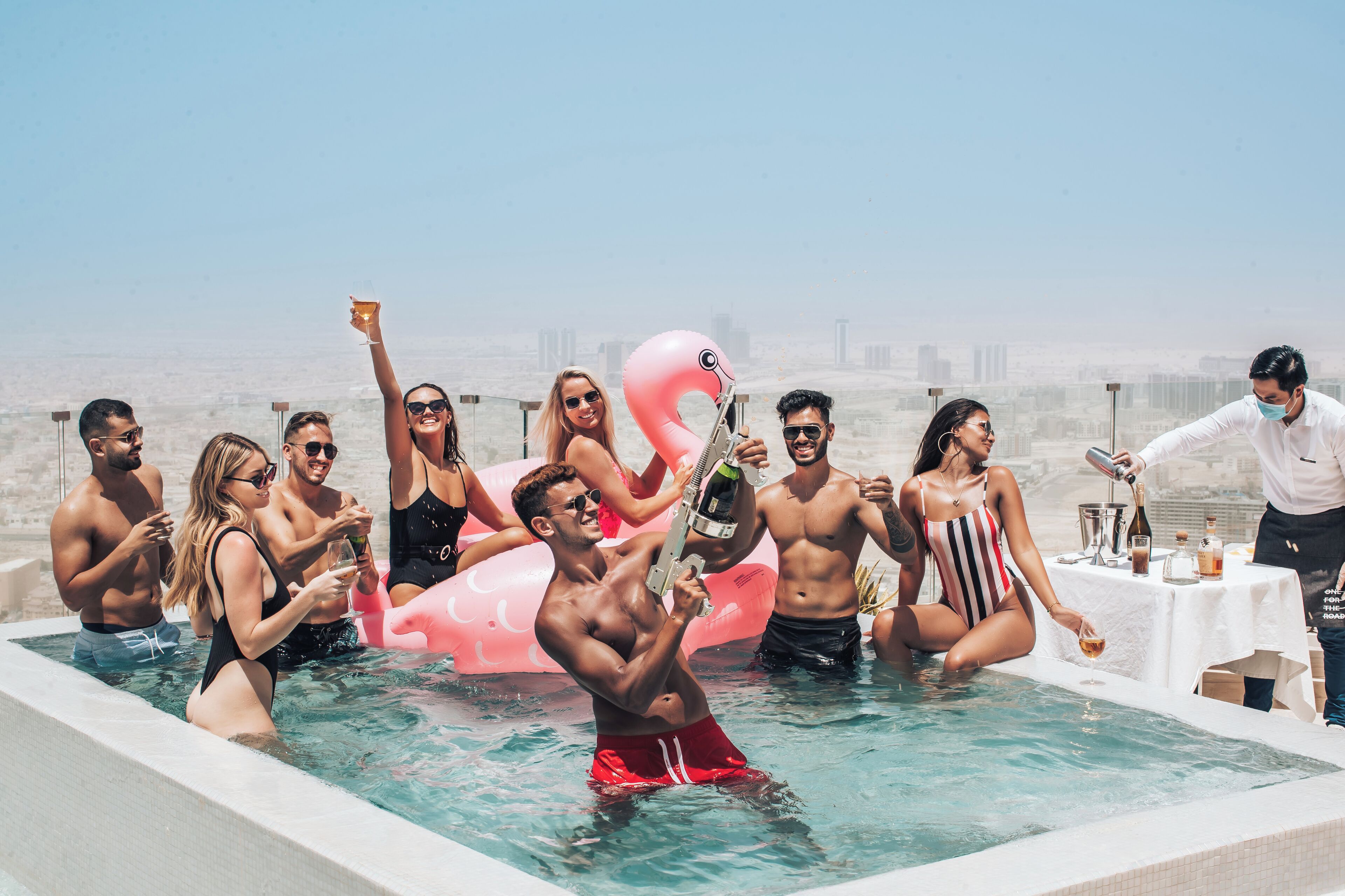 A group of people in a pool with a pink flamingo float, a table with drinks, and a cityscape in the background.