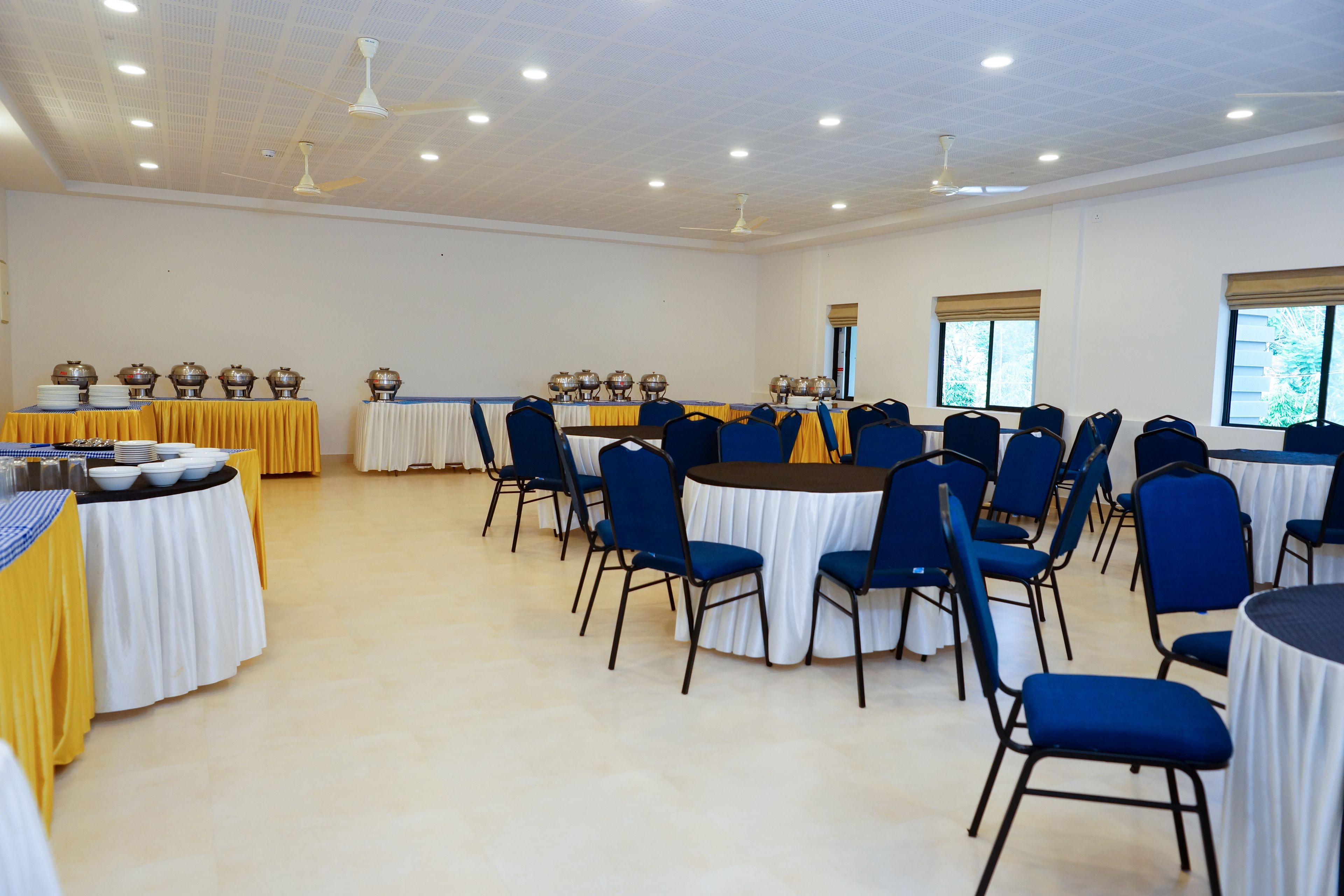 Sala de conferências com mesas, cadeiras, ventiladores de teto e janelas.