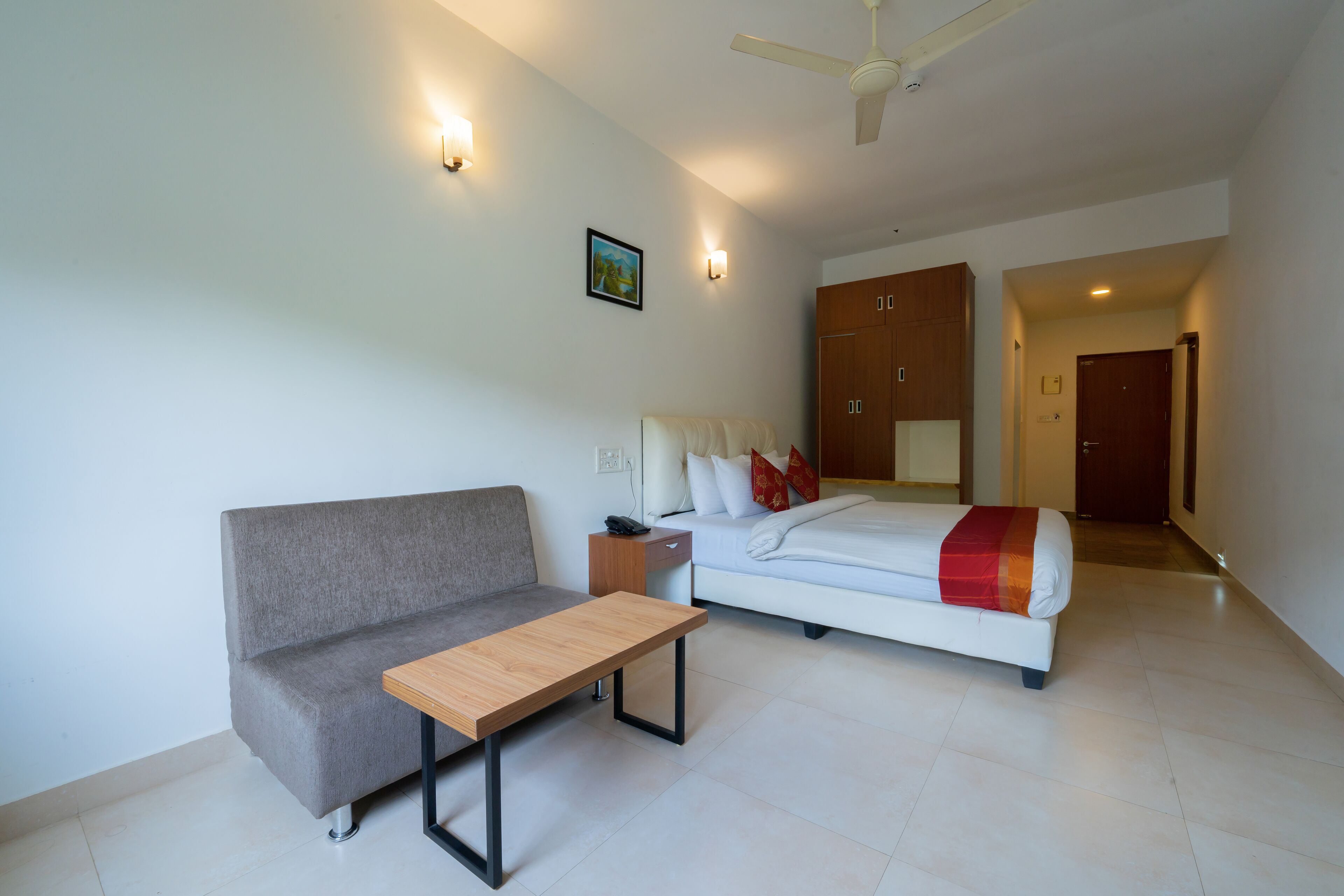 A modern hotel room with a grey sofa, a wooden table, a white bed with red and white bedding, and a ceiling fan.