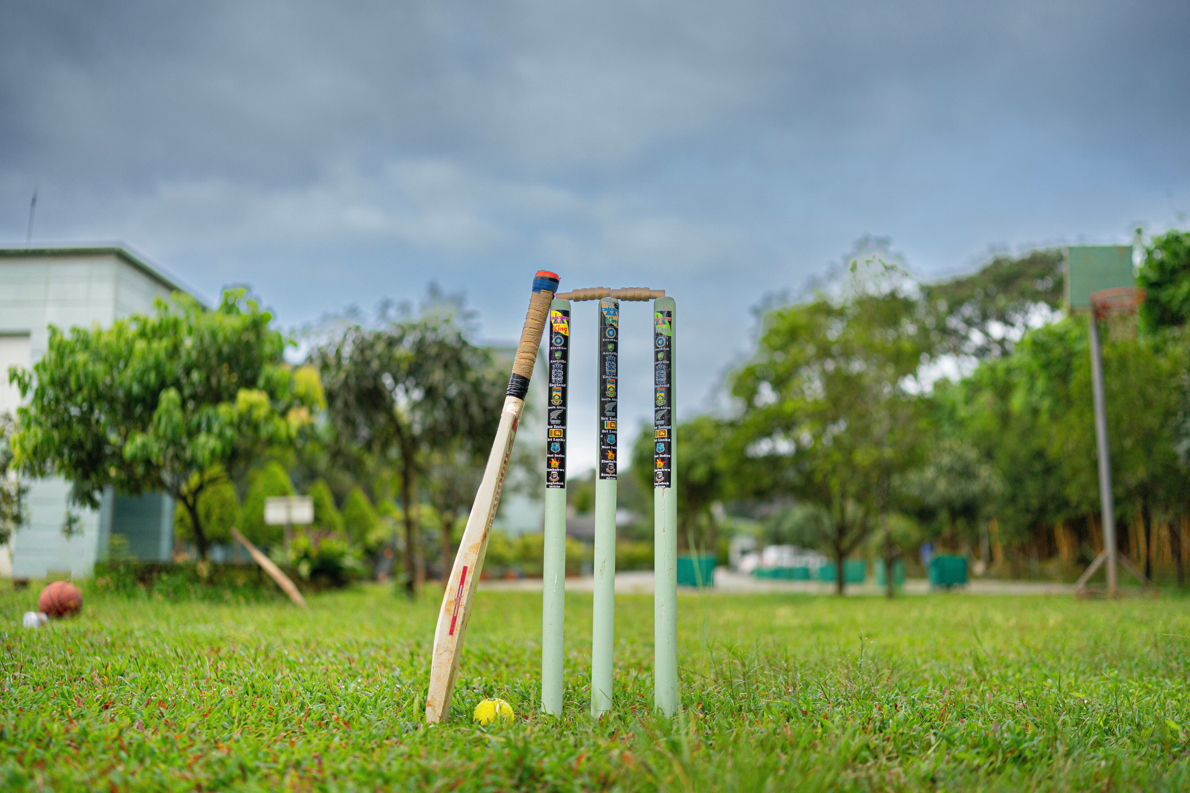 Cricket stumps and a cricket ball on a grassy field with trees and a building in the background.