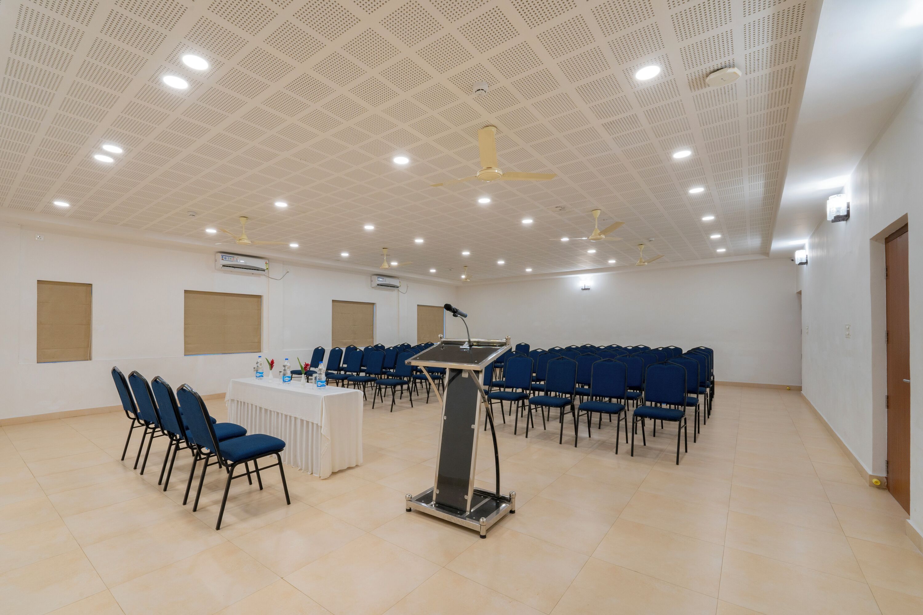 Un salón de conferencias con un atril, filas de sillas, ventiladores de techo e iluminación empotrada.