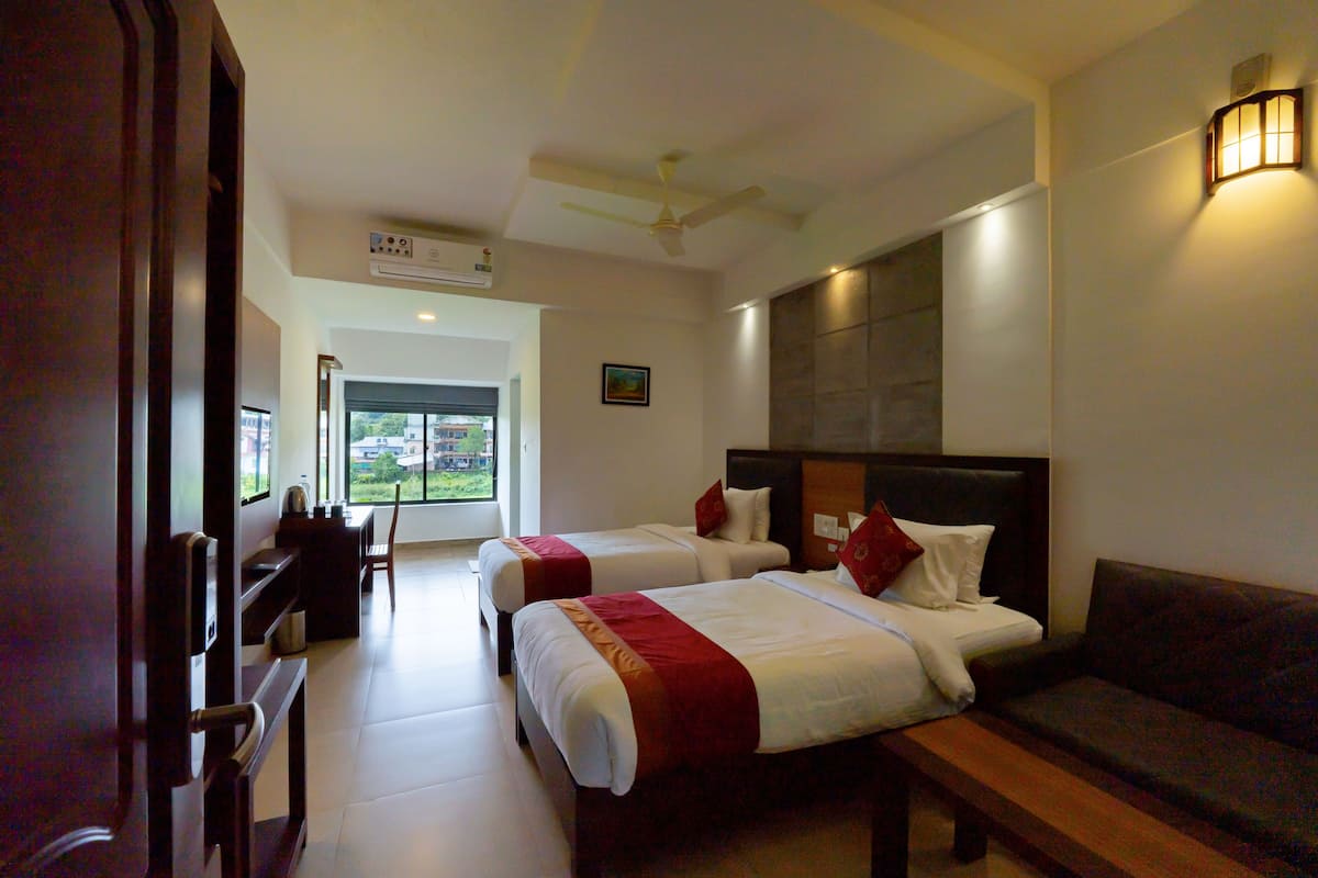 A hotel room with two beds, a bench, a sofa, a desk with a chair, and a large window.
