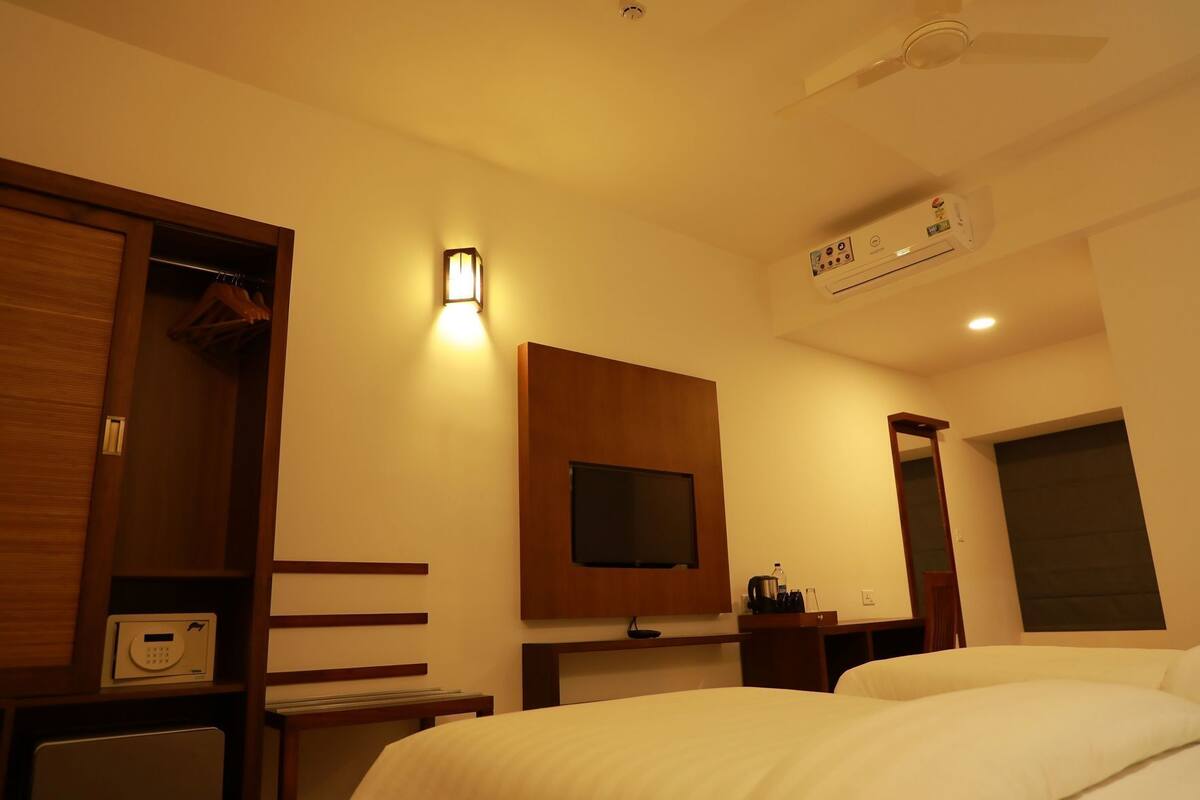 A hotel room with a bed, a television, a wardrobe, a desk with a chair, and an air conditioning unit.