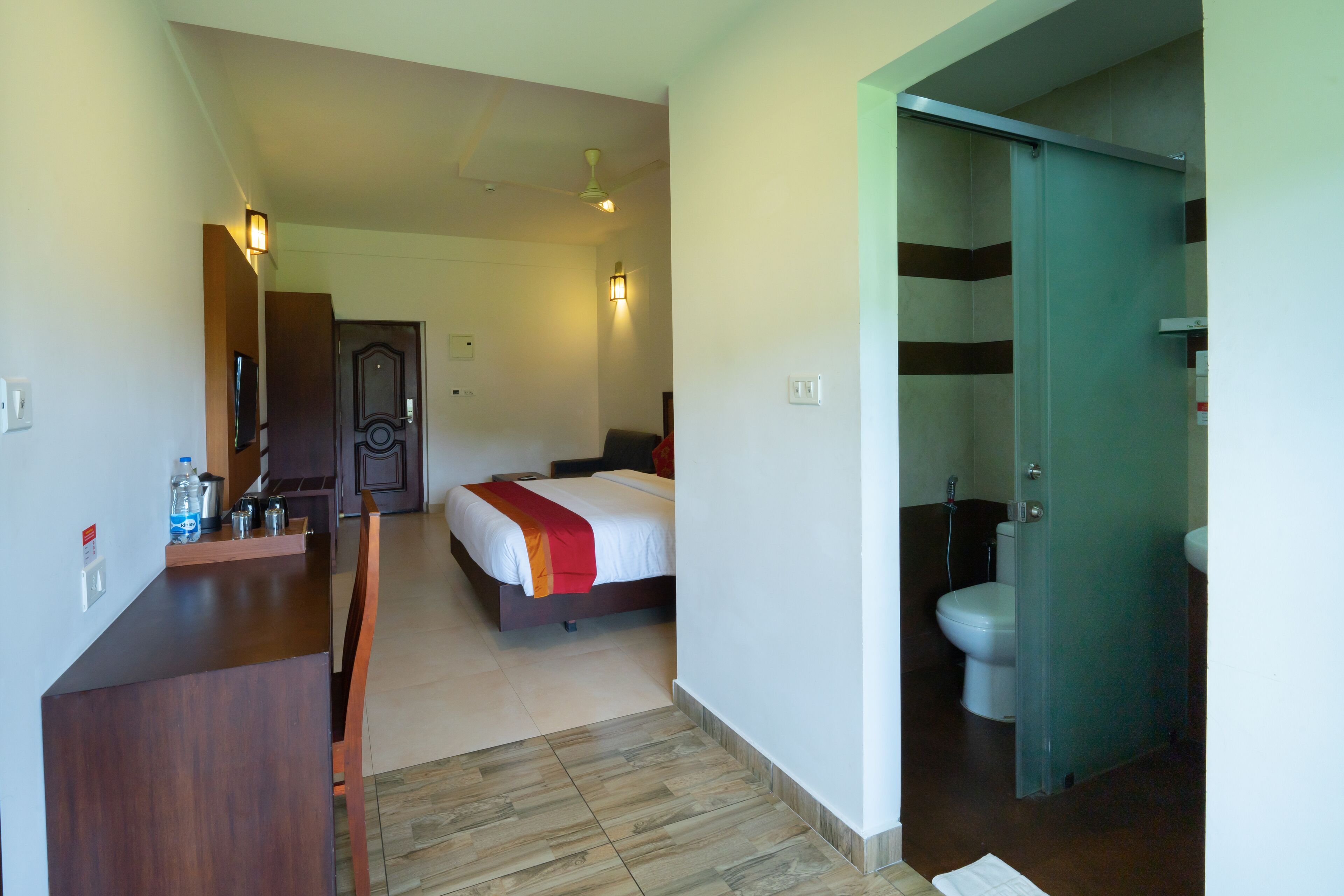 A hotel room with a bed, a desk with a chair, a bathroom with a toilet and shower, and a door leading to another room.