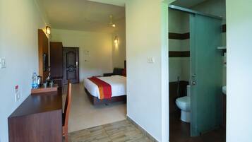Habitación de hotel con cama, escritorio con silla, baño con inodoro y ducha, y una puerta que da a otra habitación.