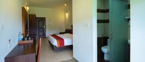A hotel room with a bed, a desk with a chair, a bathroom with a toilet and shower, and a door leading to another room.