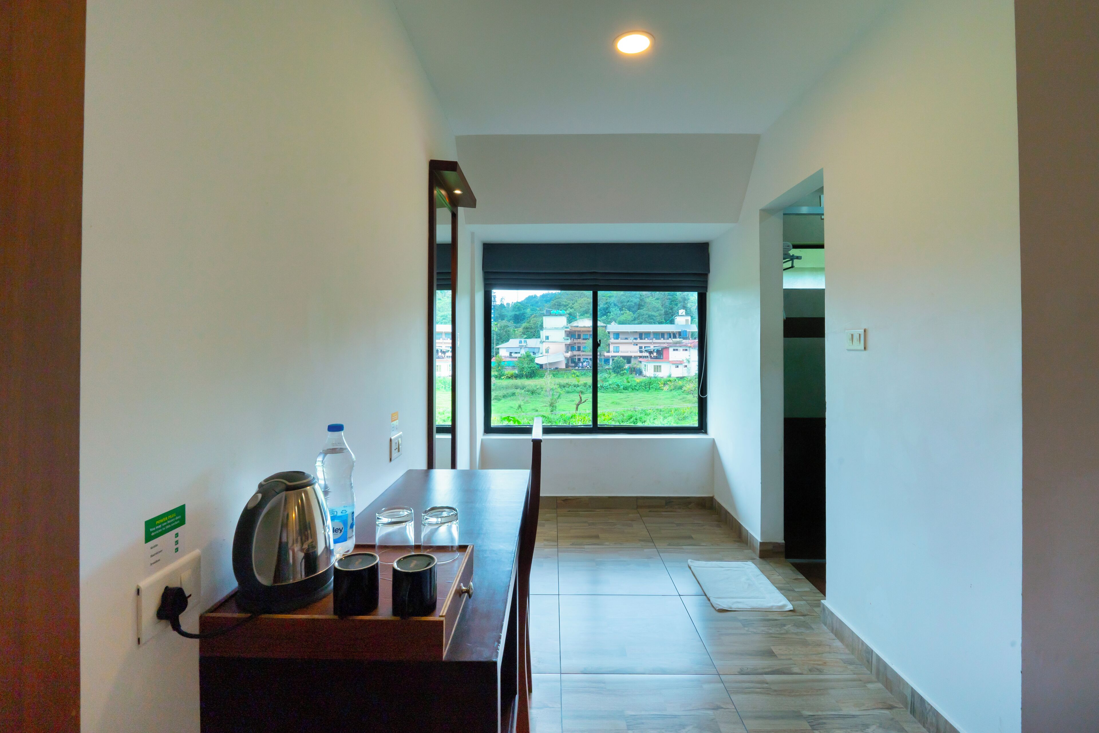 A hallway with a view of a green landscape through a large window, a kettle, a bottle, and a tray with cups.
