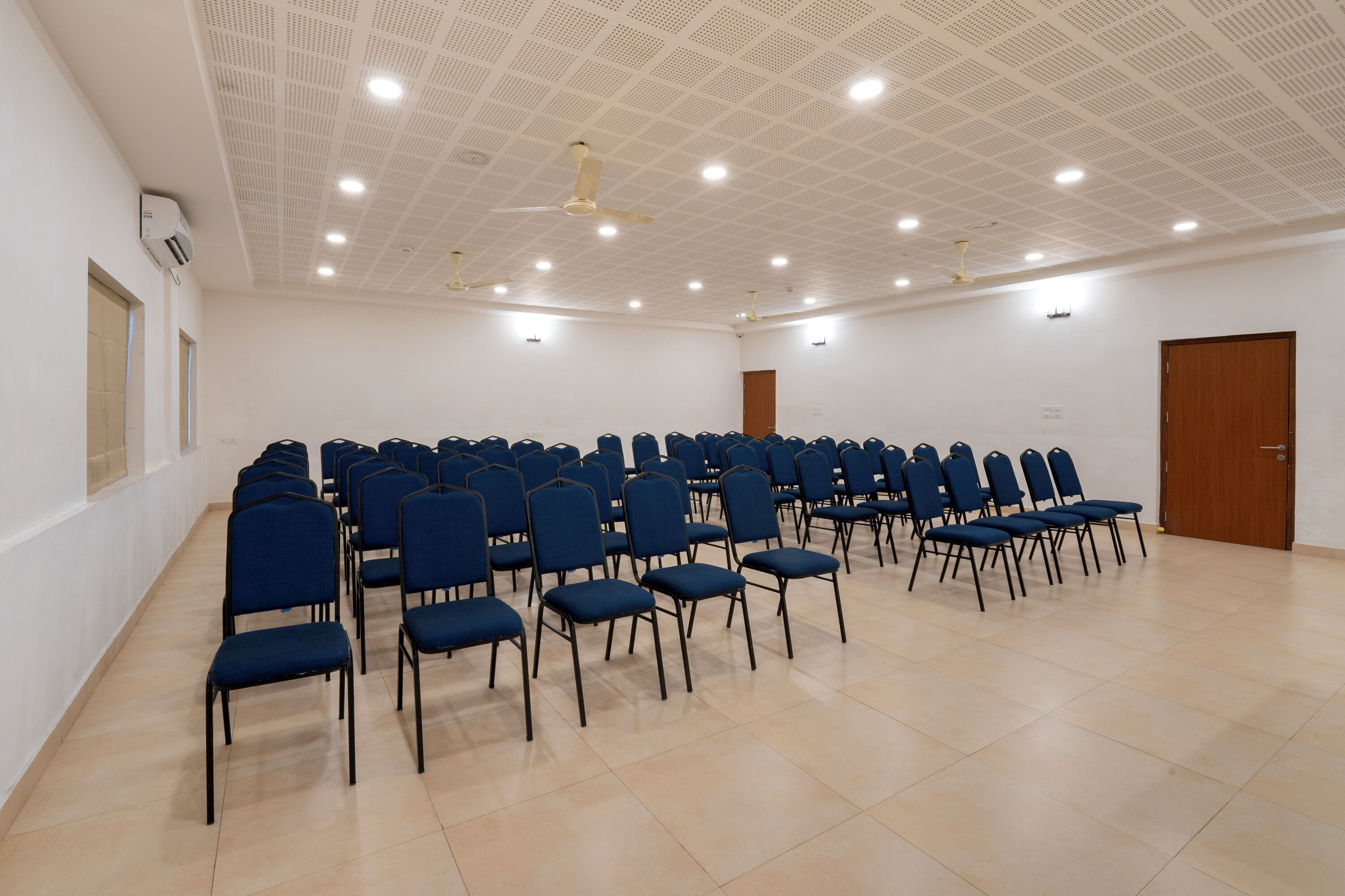 Un salón de conferencias con filas de sillas azules, un techo blanco con iluminación empotrada y una puerta de madera.