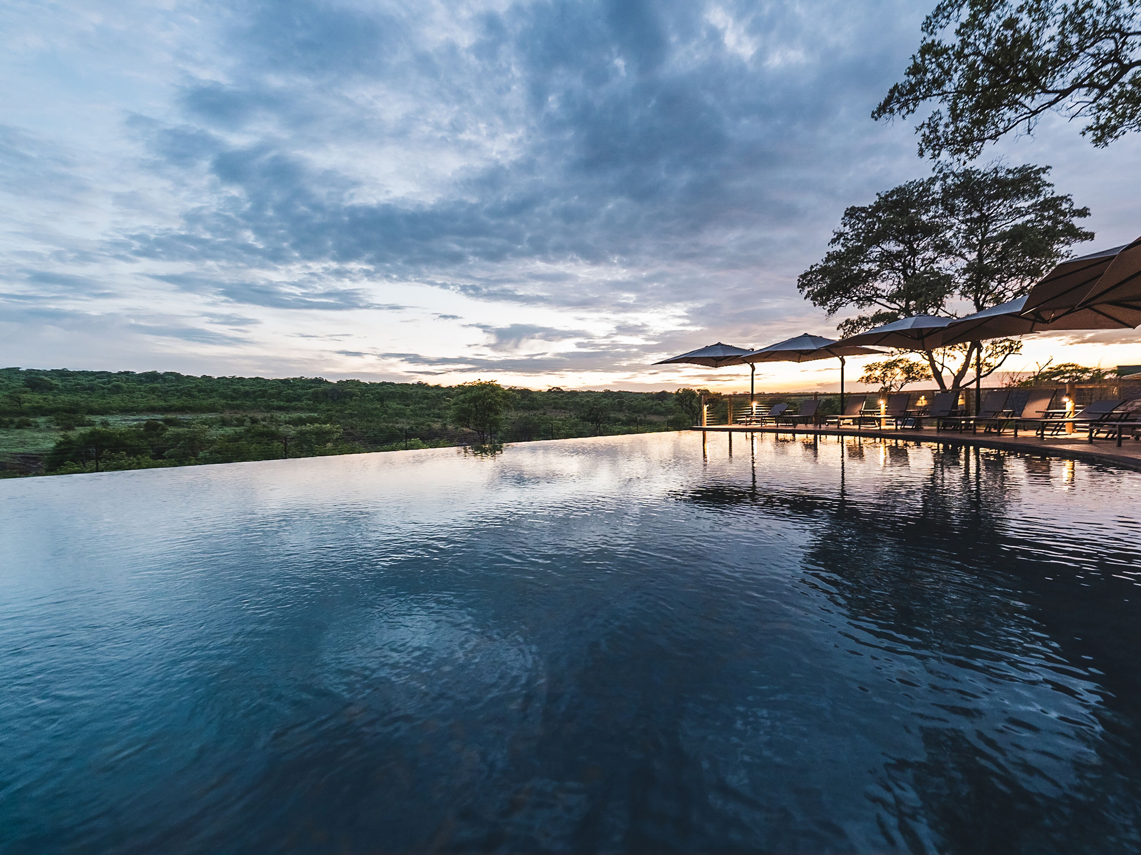 An infinity pool with a view of a tree-lined landscape and a sunset.