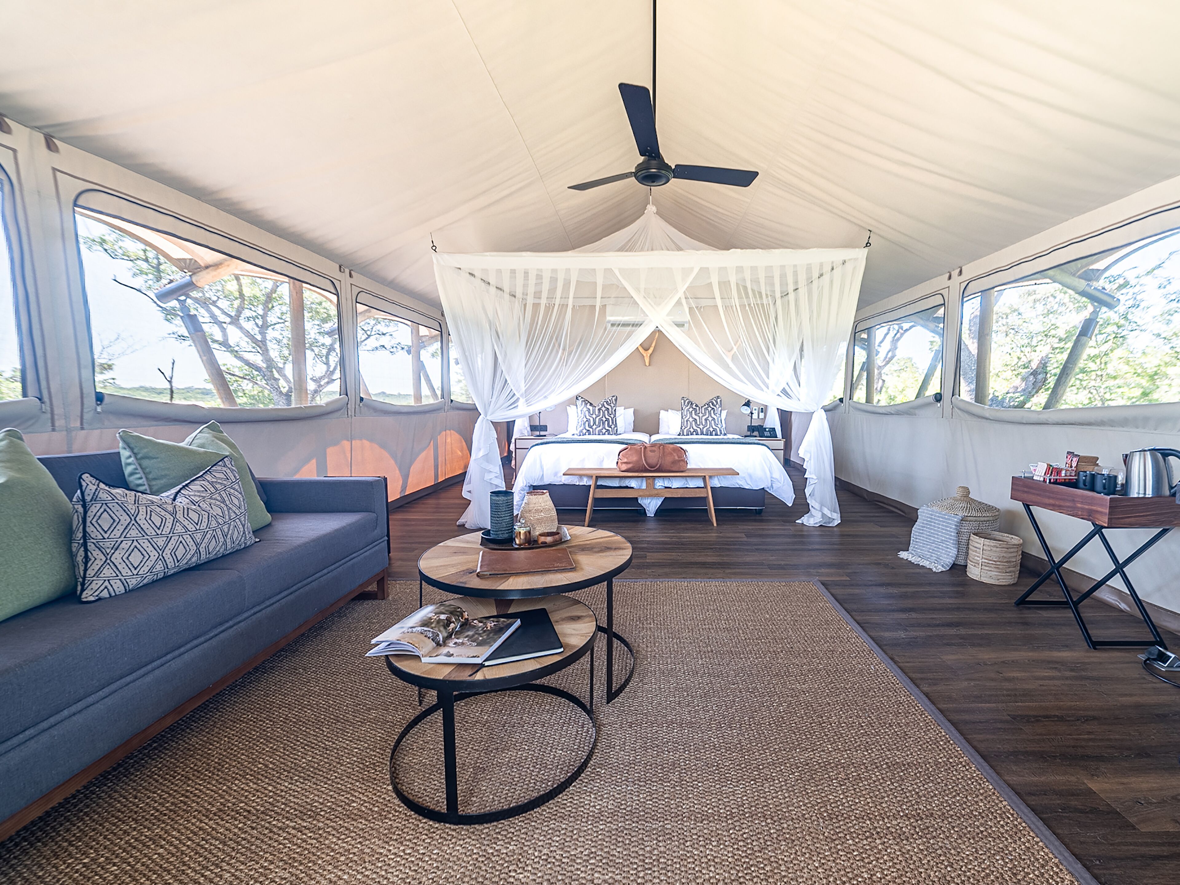 A spacious room with a large bed under a canopy, a sofa with cushions, a small table with a magazine, and a ceiling fan.