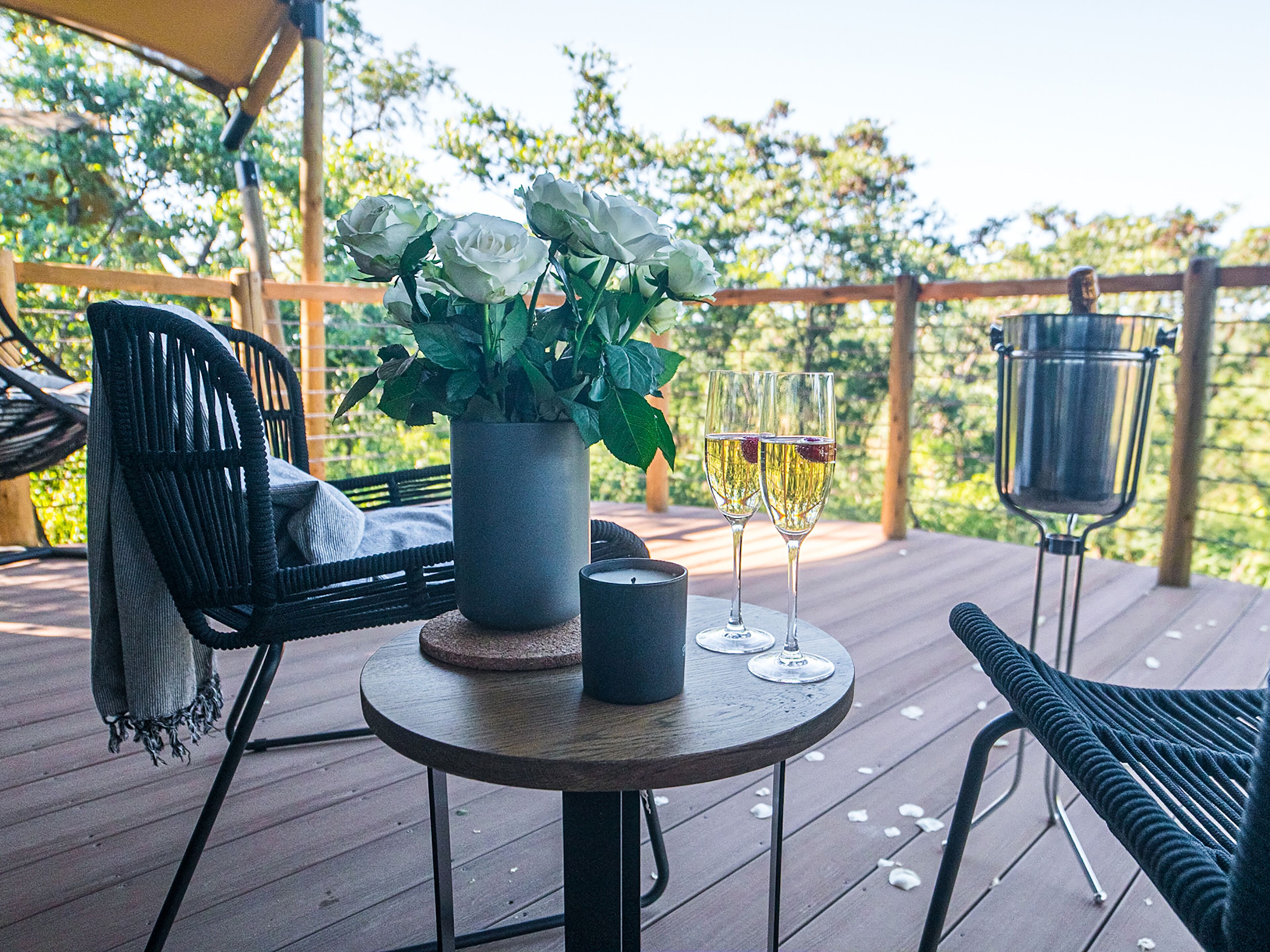 A wooden deck with a round table, two chairs, a potted plant, a candle, and two glasses of champagne.