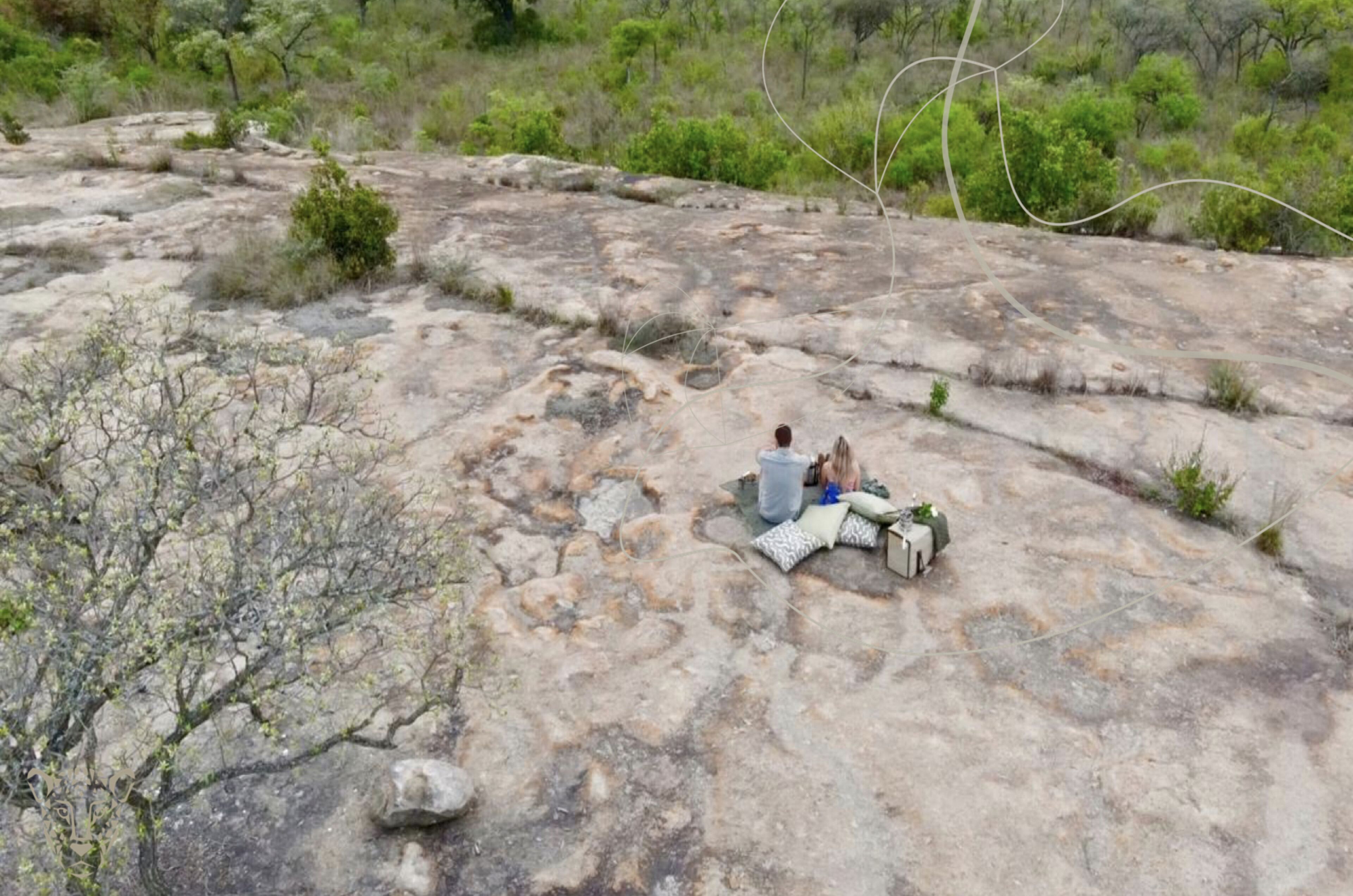 A rocky outdoor area with two people sitting on a blanket, surrounded by sparse vegetation.