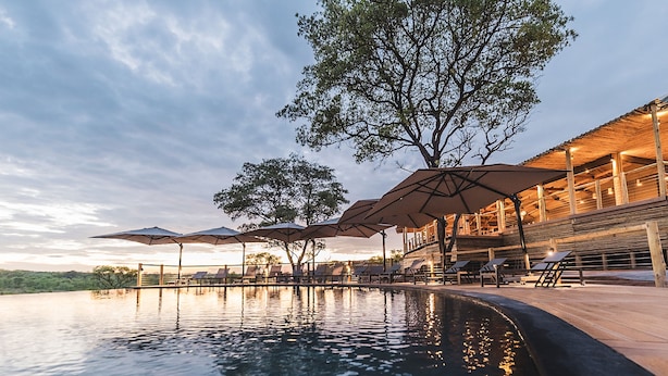Un espace aménagé au bord de la piscine, avec des parasols et des chaises longues, entouré d’arbres et bordé d’un bâtiment à la façade en bois.