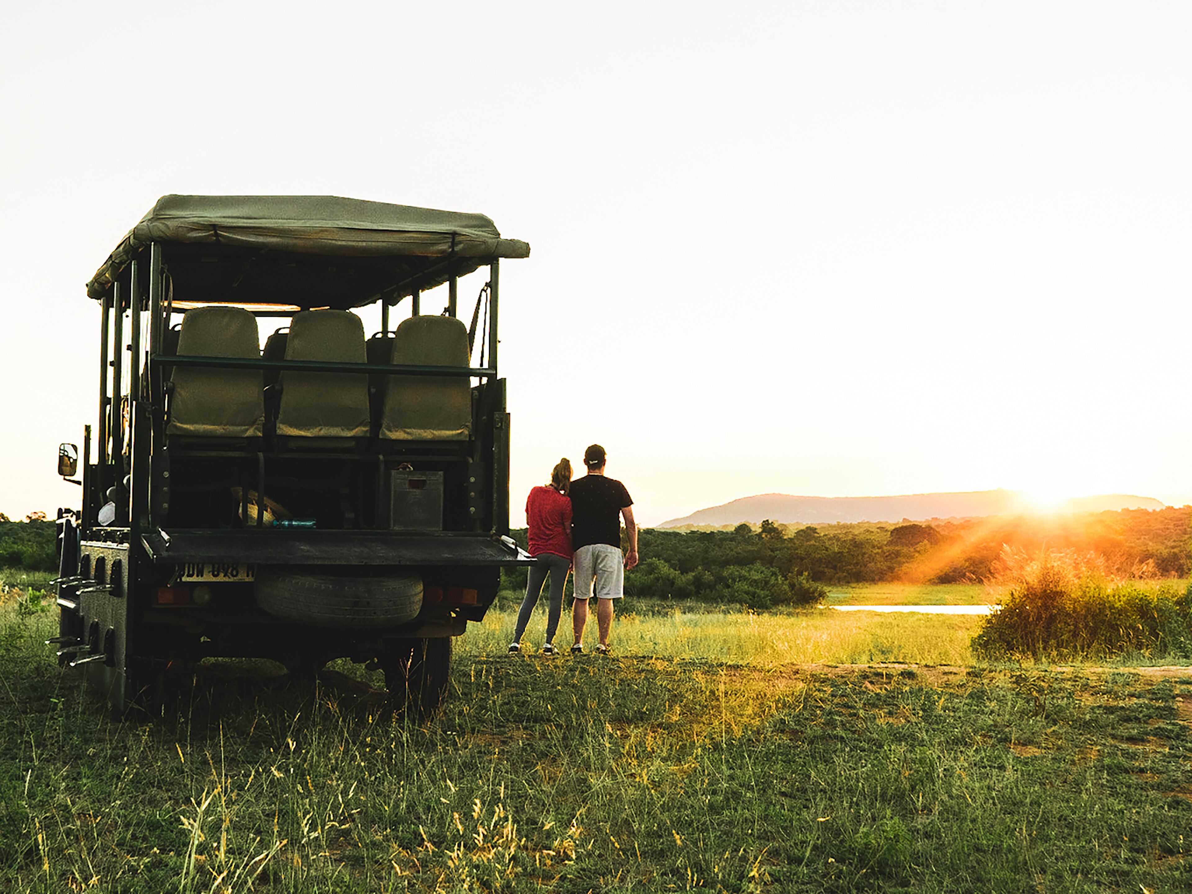 A safari vehicle with a roof and seats, parked in a grassy field with a sunset in the background.