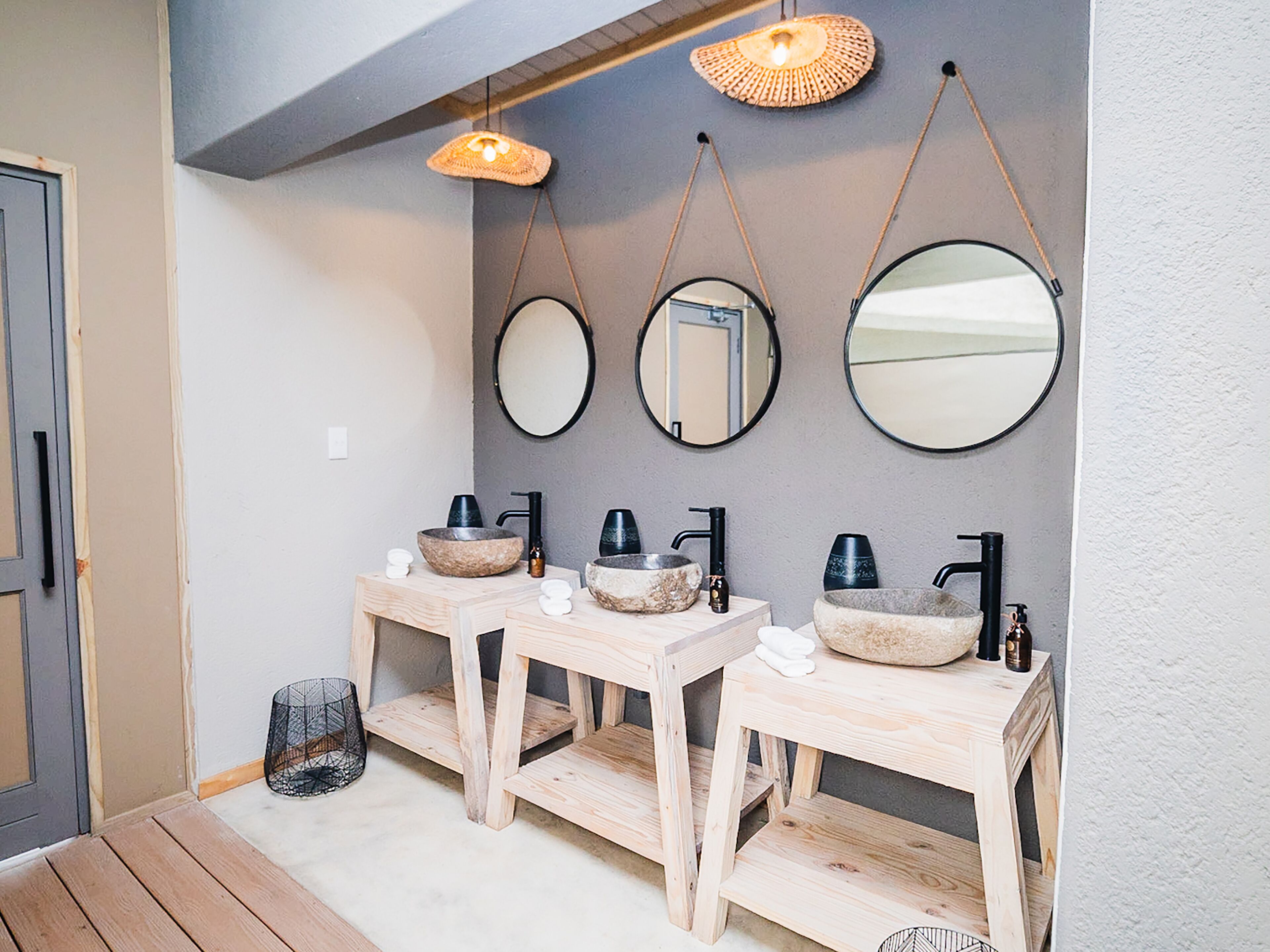 A bathroom with three wooden vanity units, each with a stone basin and black faucets. There are three wall-mounted mirrors and two hanging woven light fixtures.