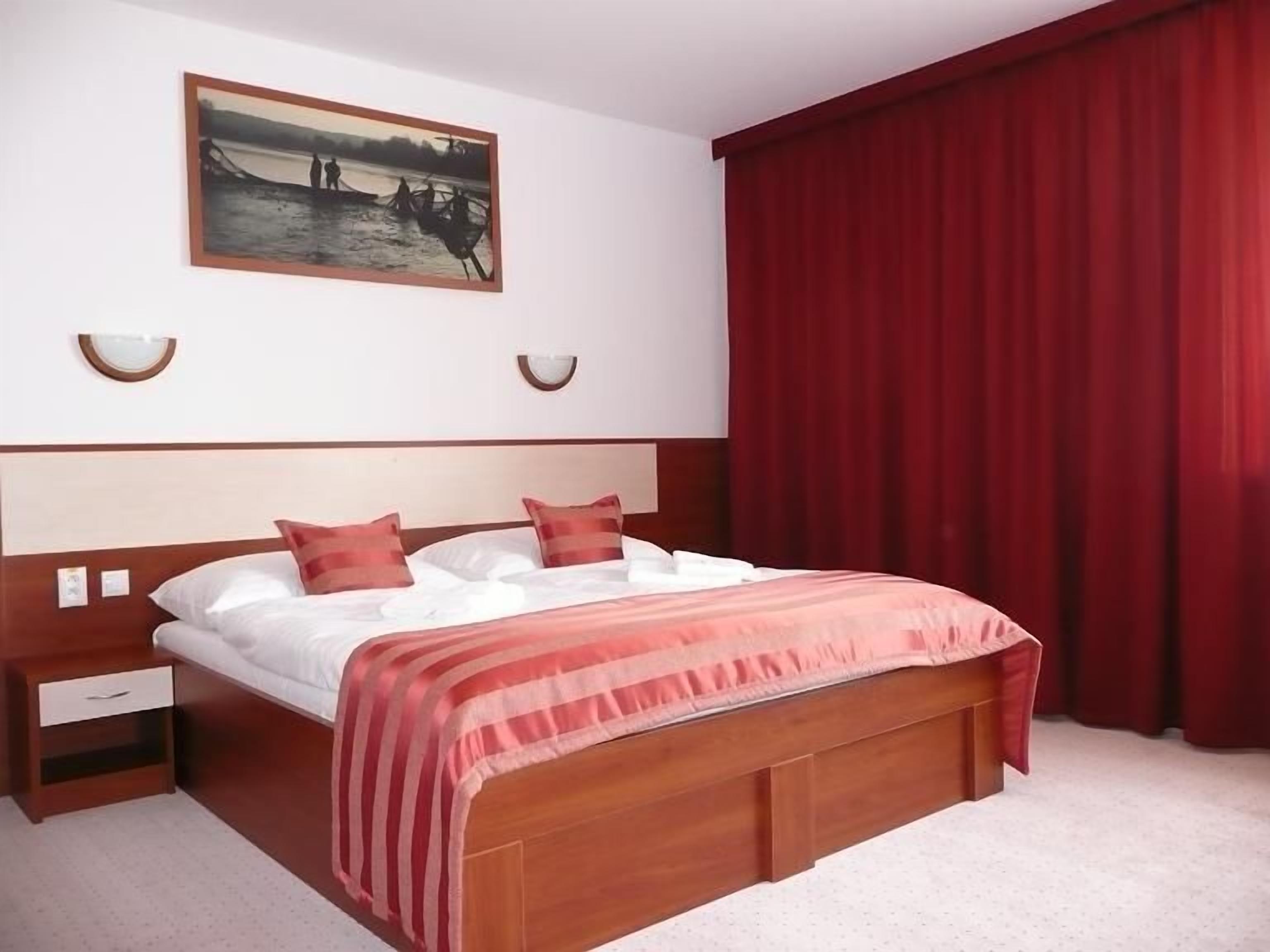 A hotel room with a large bed, a nightstand, a wooden headboard, a painting on the wall, and red curtains.
