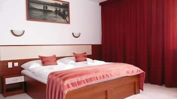 A hotel room with a large bed, a nightstand, a wooden headboard, a painting on the wall, and red curtains.