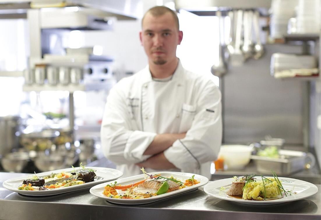 Un chef se tient derrière un comptoir, avec trois assiettes contenant des plats préparés, dans une cuisine professionnelle.