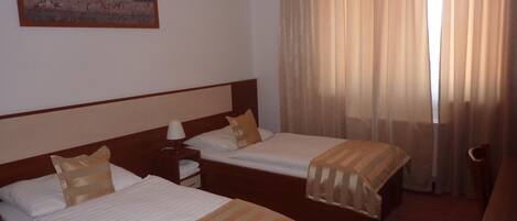 Habitación de hotel con dos camas, una mesita de noche, una lámpara, un cuadro de un paisaje boscoso y un ventanal con cortinas.