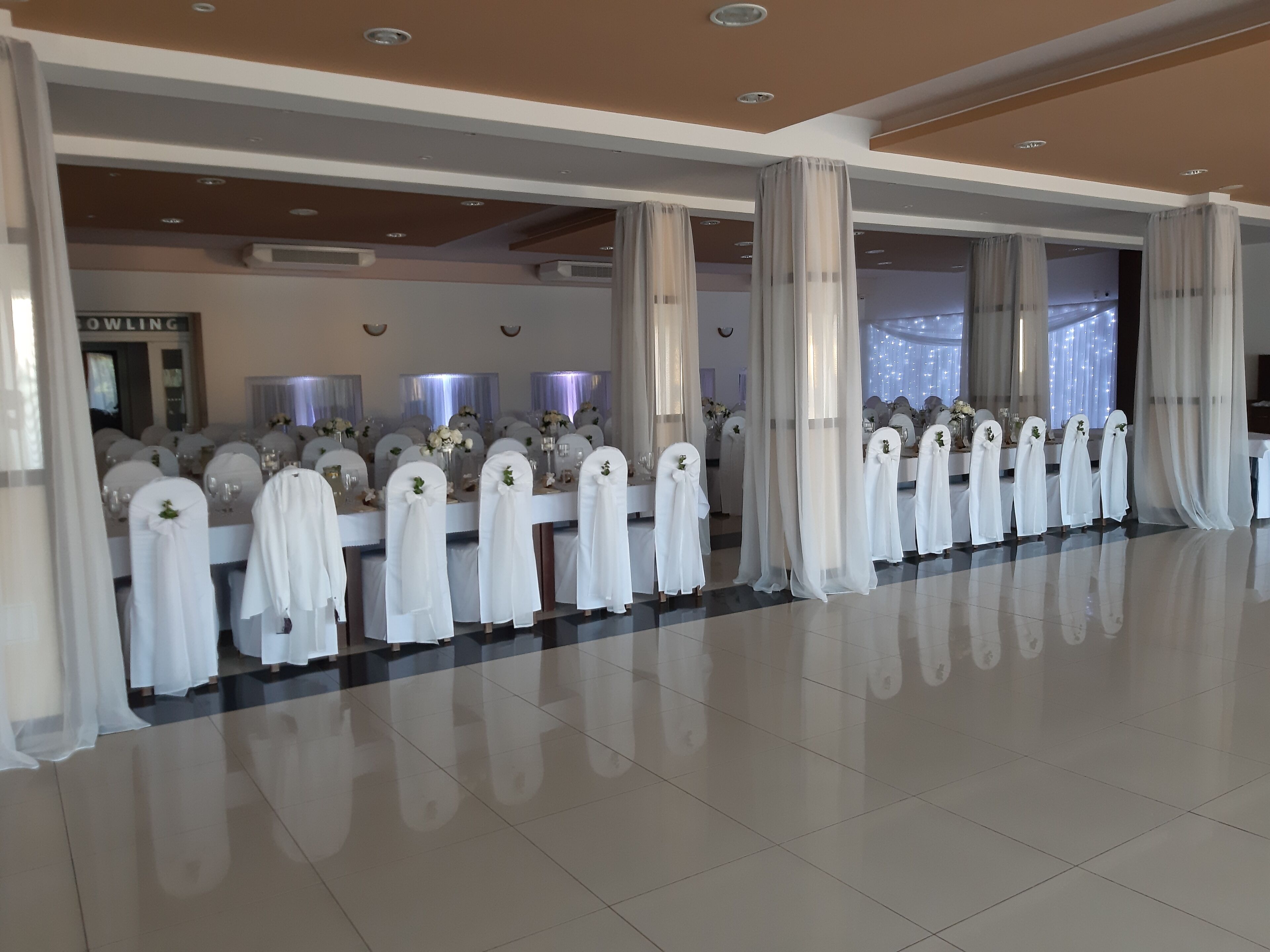 A spacious hotel lobby with rows of white-covered chairs and tables, large windows, and a modern ceiling design.