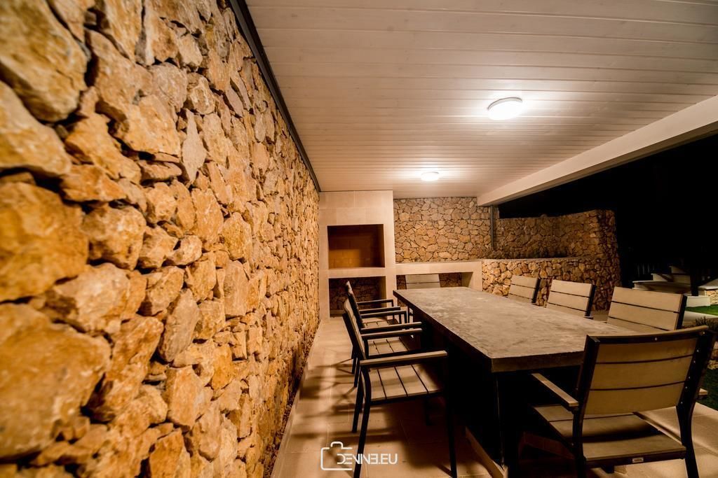 A stone-walled dining area with a long table and chairs, recessed ceiling lights, and a stone fireplace.