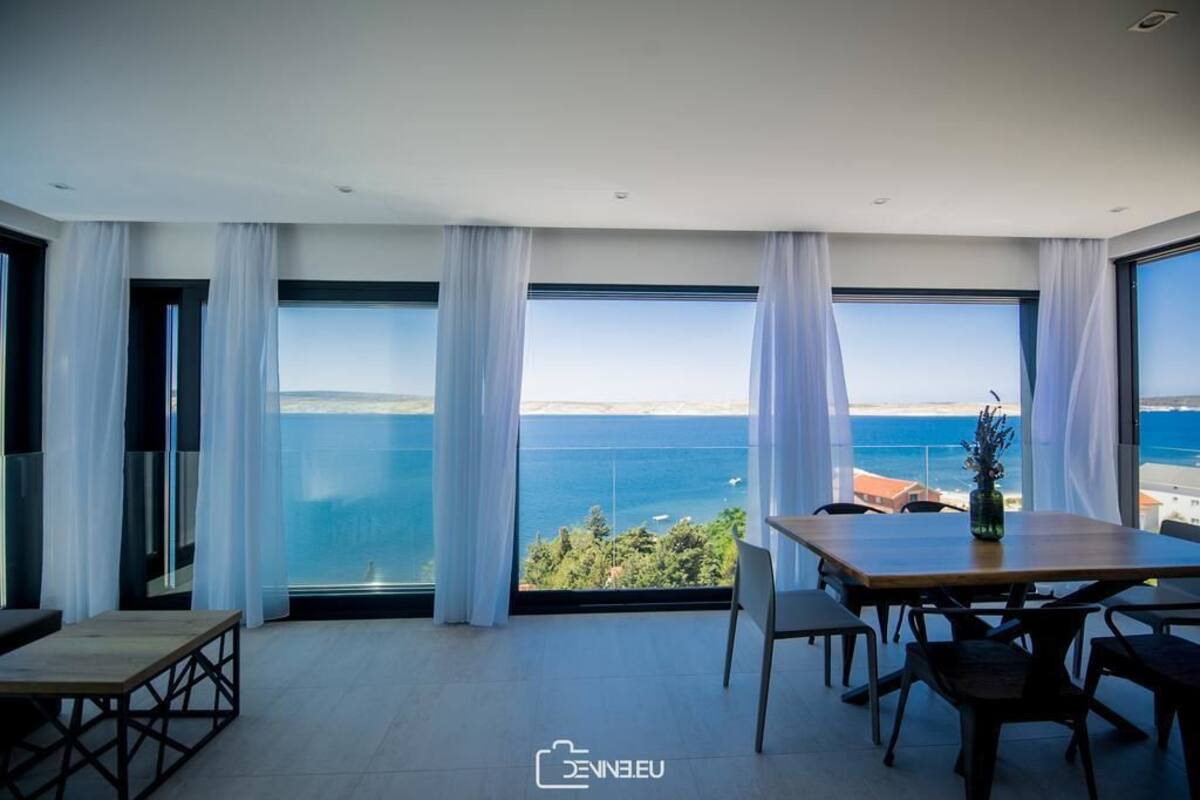 A modern room with a large window offering a view of the sea and a distant landscape. There is a wooden table with chairs and a bench.