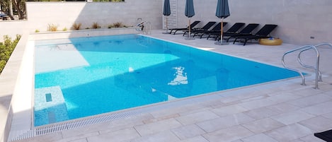 A rectangular swimming pool with a tiled edge and a metal ladder. There are lounge chairs and umbrellas along the poolside.