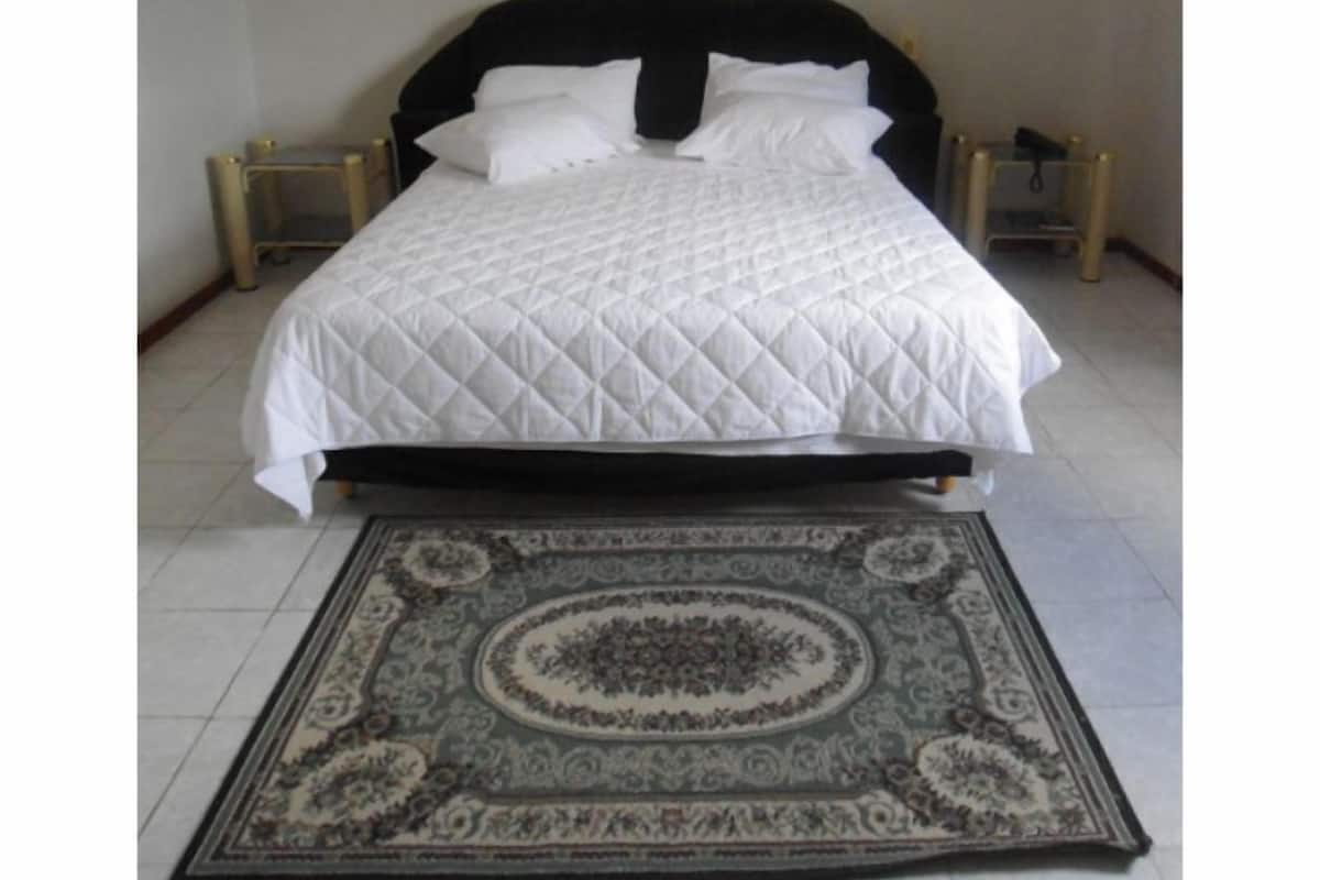 Ein Bett mit schwarzem Kopfteil und weißer Bettwäsche, ein gemustertes Teppich und ein an der Wand hängendes Kunstwerk.