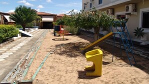 A sandy courtyard with a playground, including a slide and climbing structures, surrounded by buildings with air conditioning units. - Hotel Sun Valley (Senhor Do Bonfim)