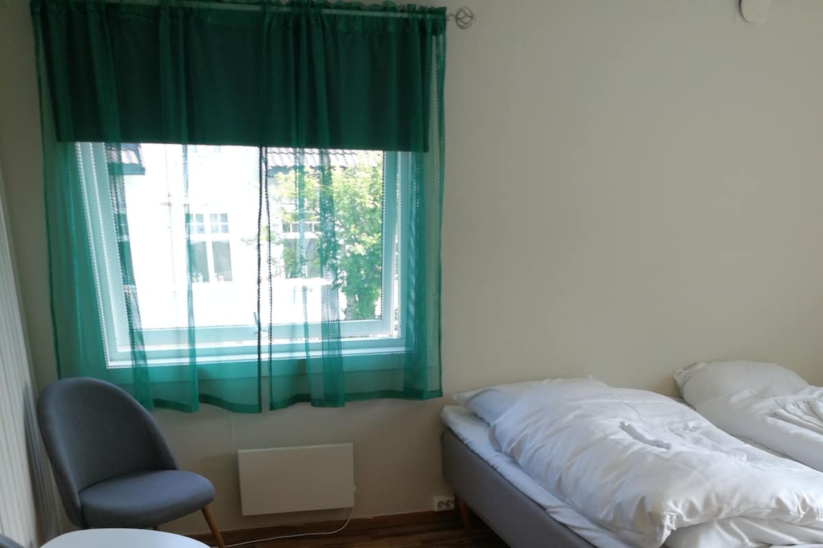 Una habitación pequeña y ordenada con una cama, dos sillas, un radiador y una ventana con cortina verde.