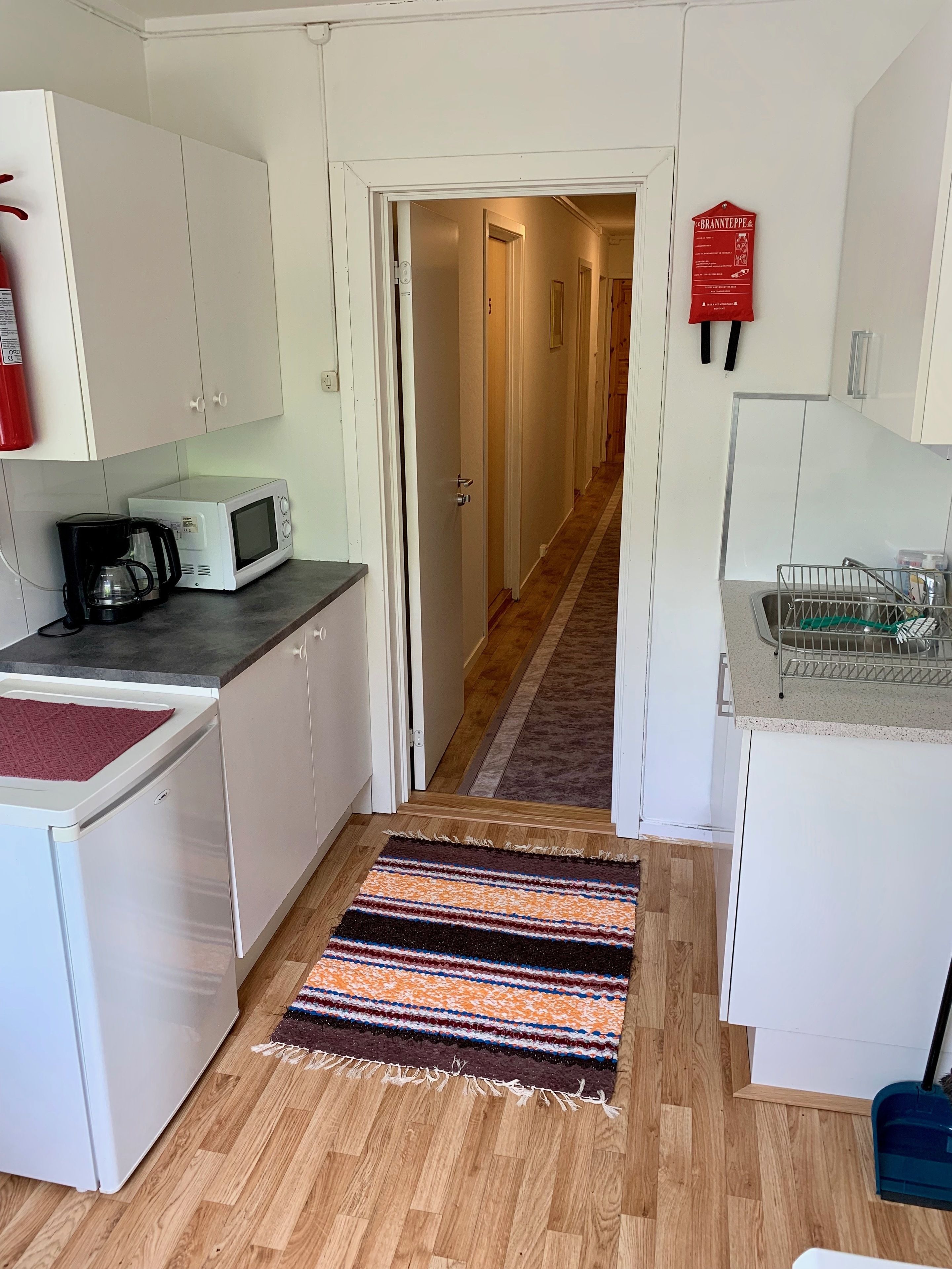A compact kitchen with white cabinets, a microwave, a coffee maker, and a red fire extinguisher. A striped rug leads to a hallway with a door marked '5'.