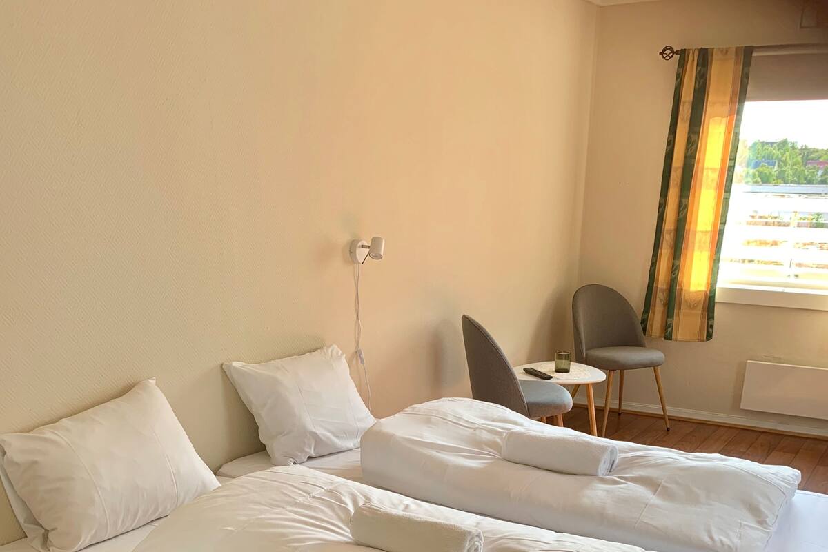 Habitación de hotel con dos camas, una mesita, una silla y un ventanal con cortinas.