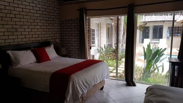 Un dormitorio con pared de ladrillo, cama con ropa de cama blanca y roja, y vistas a un patio con plantas y piscina.