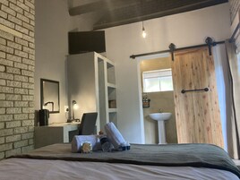 A hotel room with a bed, a desk with a computer, a chair, a small table with toiletries, and a bathroom with a sink and window.