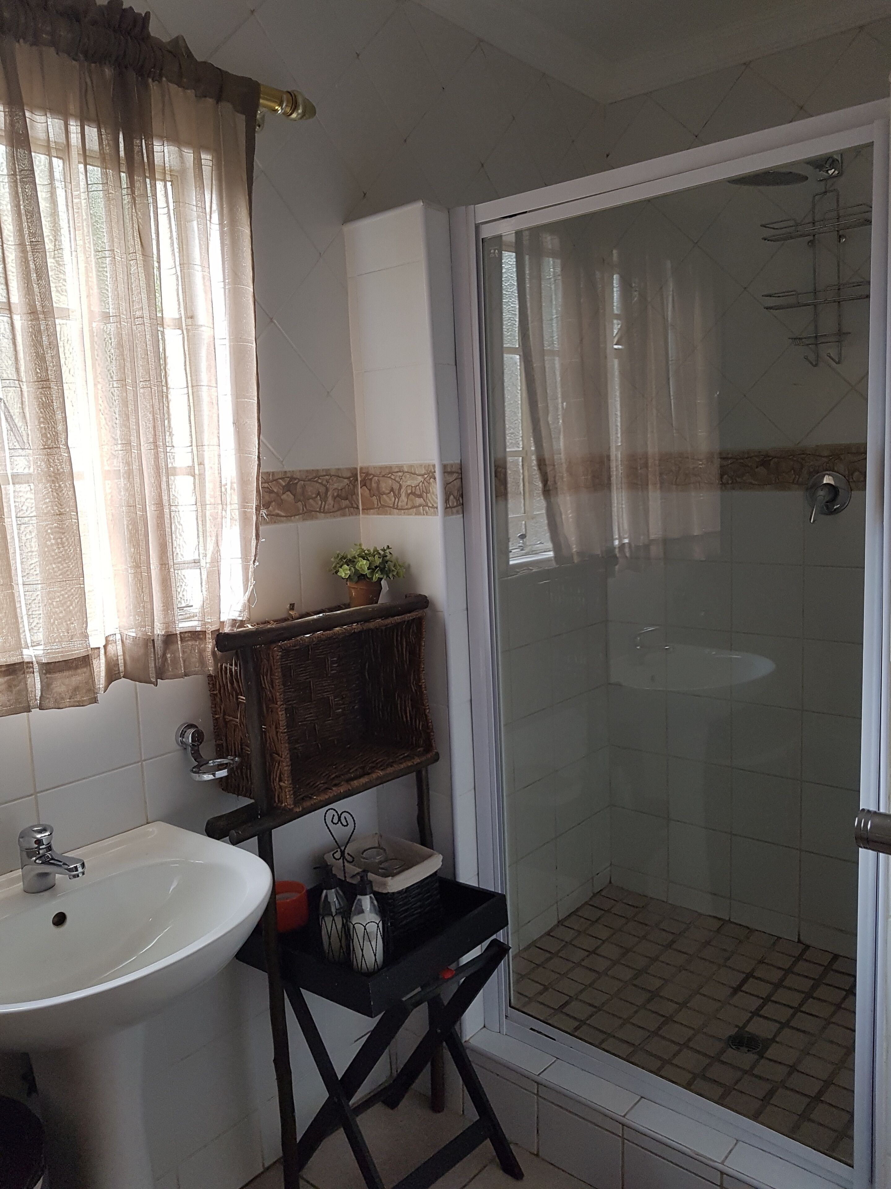 A bathroom with a sink, a mirror, a shower with a glass door, and a window with sheer curtains.