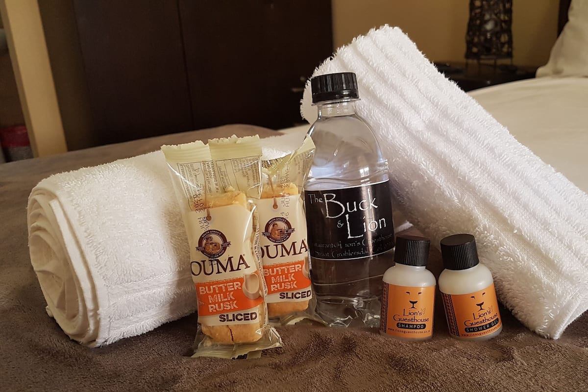 A hotel room with a bed, a white towel, and toiletries including OUMA butter milk rusks, Buck & Lion shower gel, and a bottle of water.