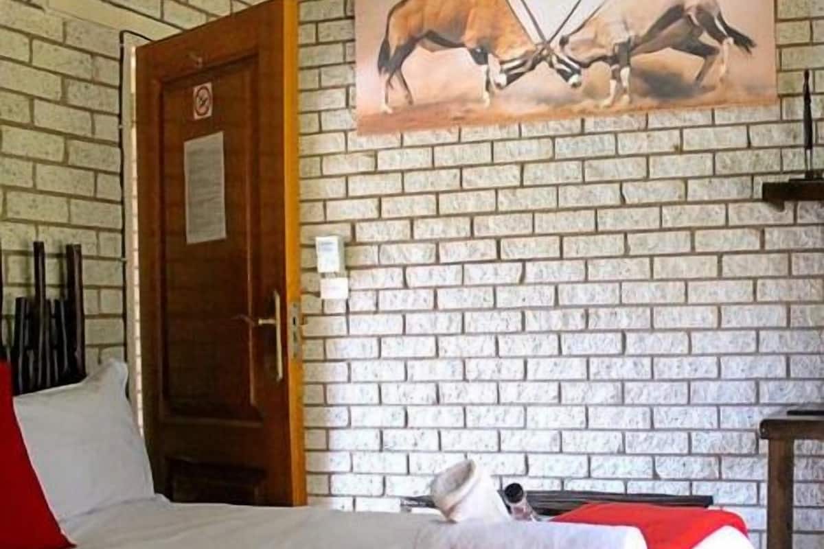 A room with a brick wall, a wooden door, a bed with a red blanket, and a painting of two antelopes.