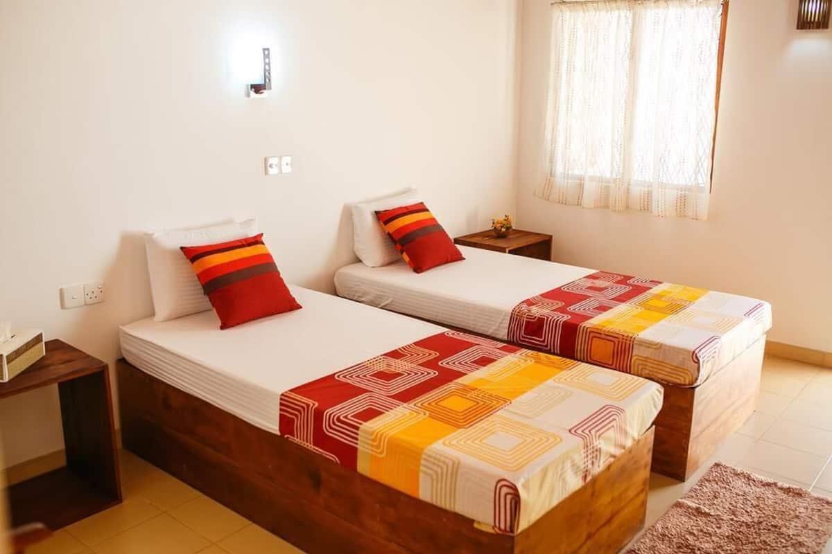 A double room with two beds, each with a patterned bedspread and pillows, a small bedside table, and a window with sheer curtains.