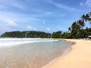 A beach with clear water, palm trees, and a distant forested area. - Hostel First Mirissa (Mirissa)