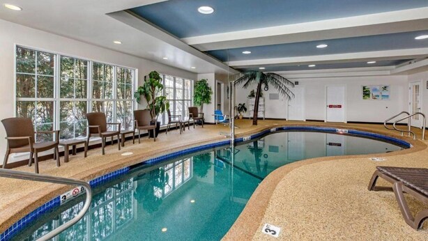 Une piscine intérieure, agrémentée de chaises et de plantes en pot, entourée de fenêtres et surmontée d’un plafond bleu.