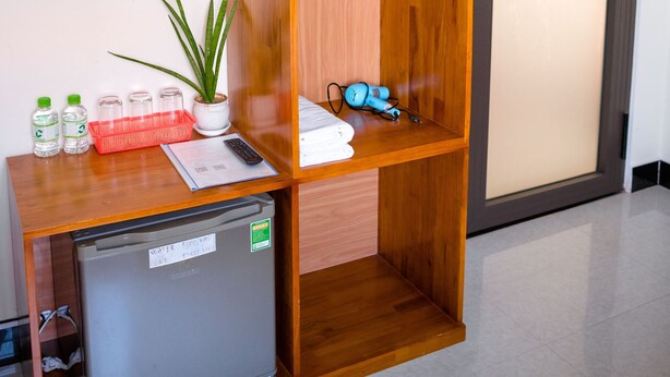 Una habitación compacta con un armario de madera, un refrigerador pequeño, un escritorio con una planta y una puerta con lámpara.