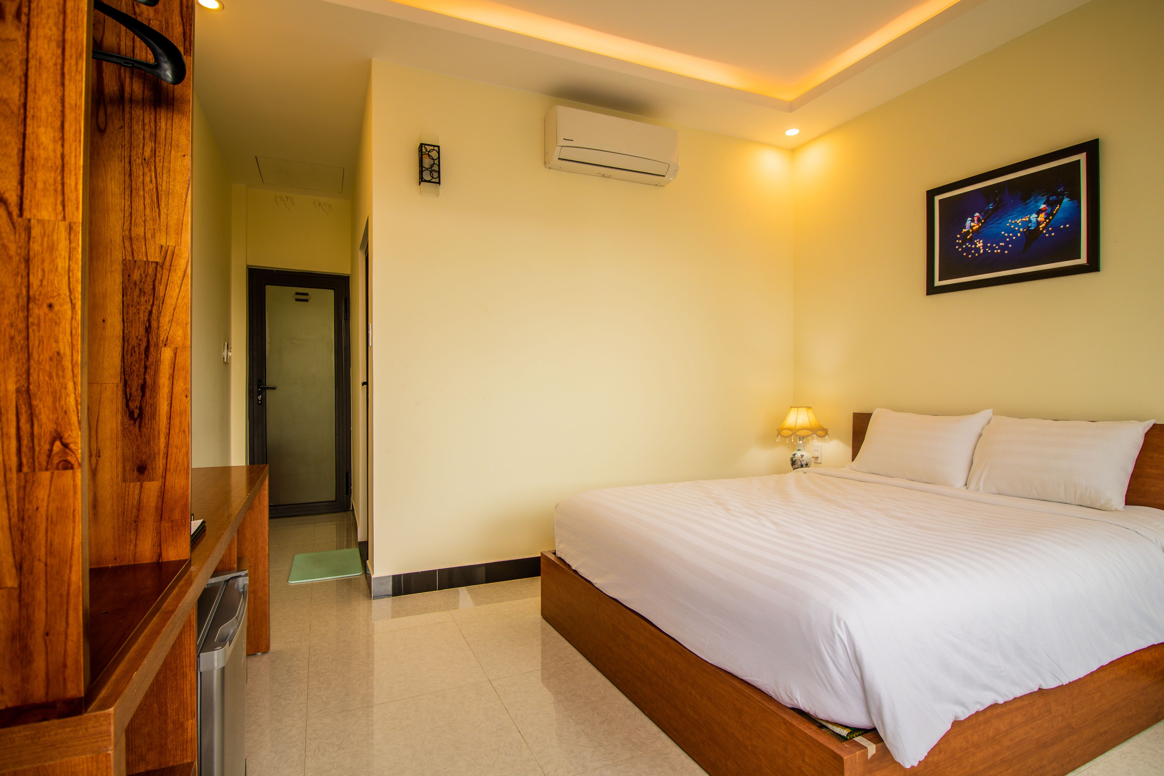 A hotel room with a wooden bed frame, a white bedspread, a wall-mounted air conditioner, a framed picture, and a wooden bench.