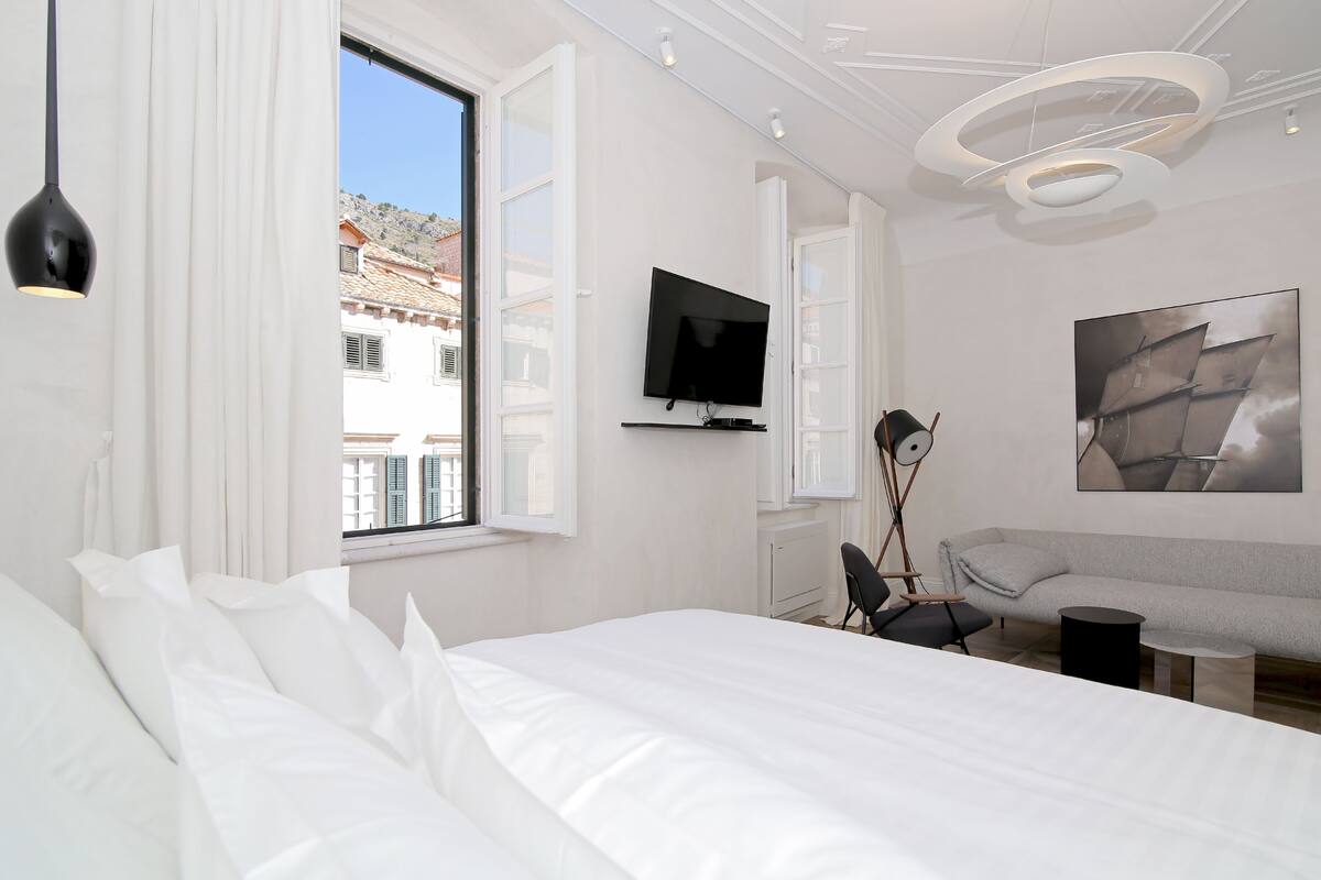 A modern hotel room with a large bed, a TV mounted on the wall, a sofa, and a view of buildings outside the window.