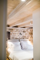 Quarto com parede de pedra, vigas de madeira no teto e roupa de cama branca.