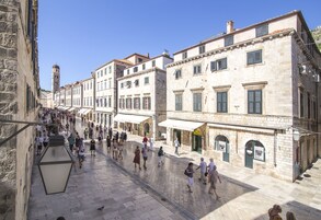 Uma rua histórica com edifícios de pedra, céu claro e pedestres.